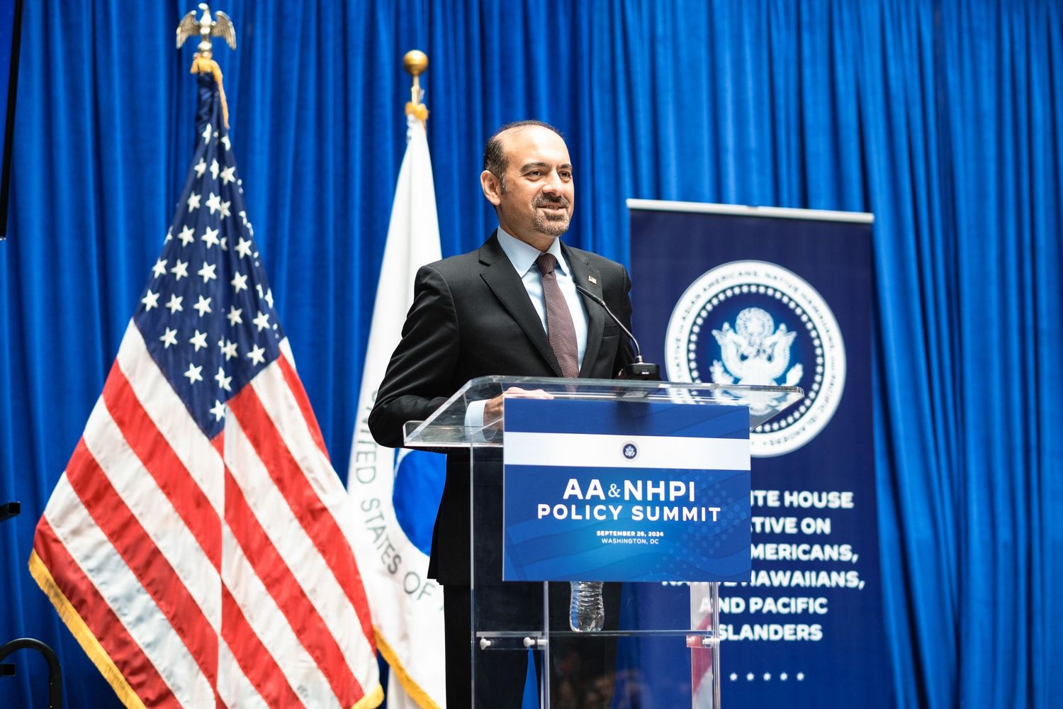 Man speaking at podium during AA&NHPI Policy Summit with US flag backdrop at White House initiative event.