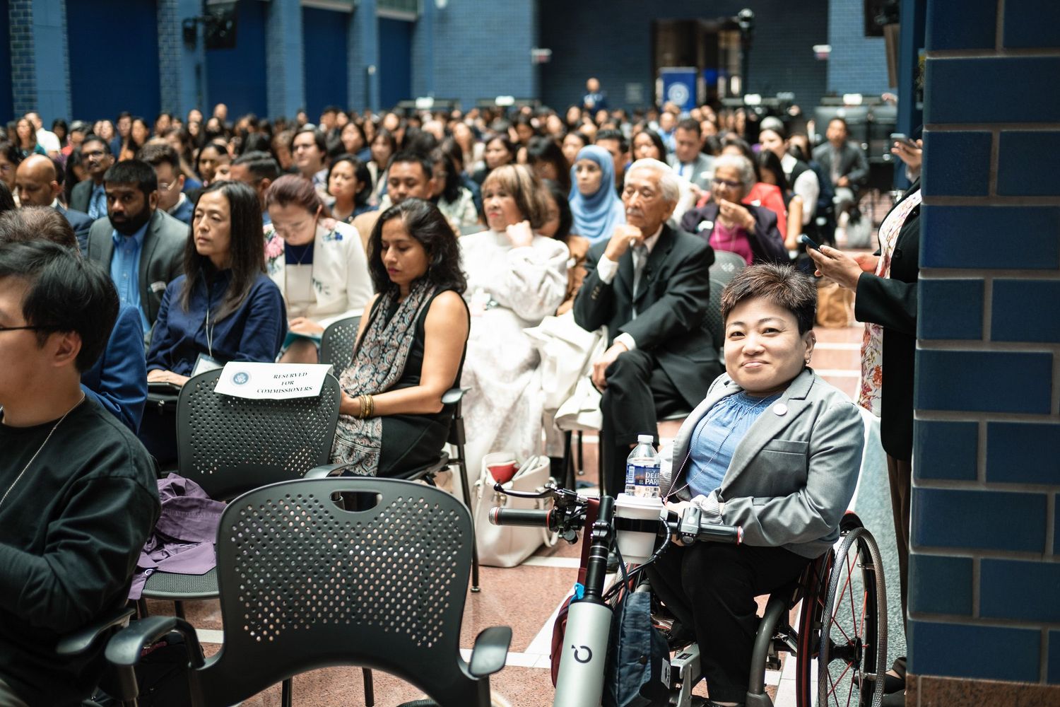 Diverse audience seated in auditorium, including a woman in a wheelchair, attending a large indoor event or conference.