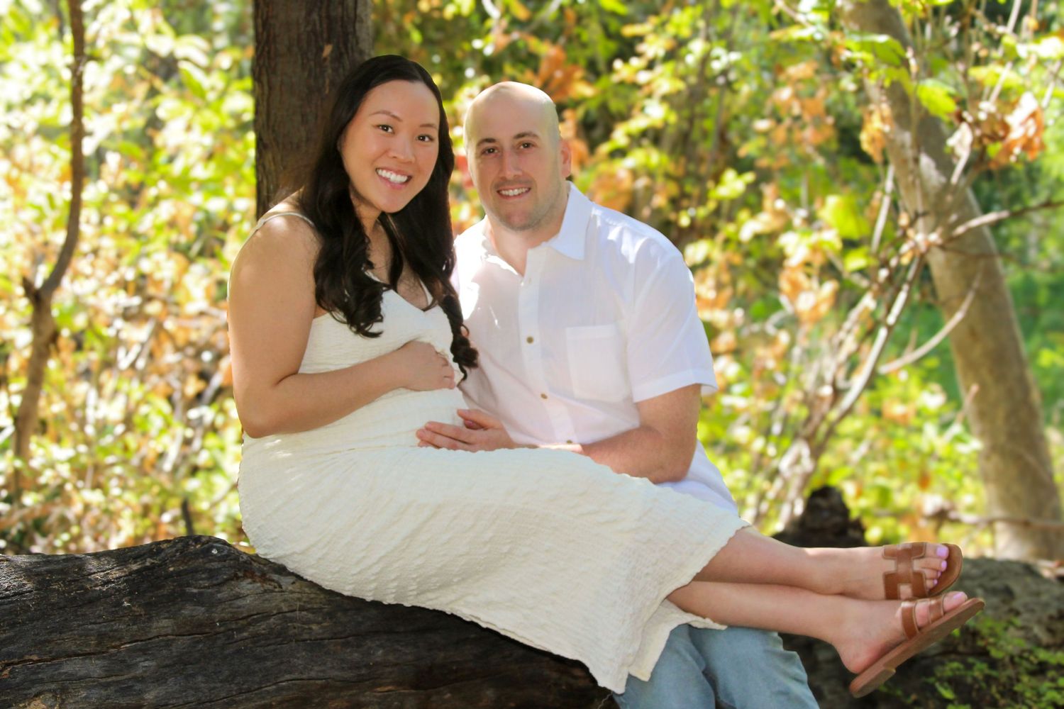 Smiling couple sitting on a log in a lush green forest, woman in white dress, man in white shirt, outdoor portrait.