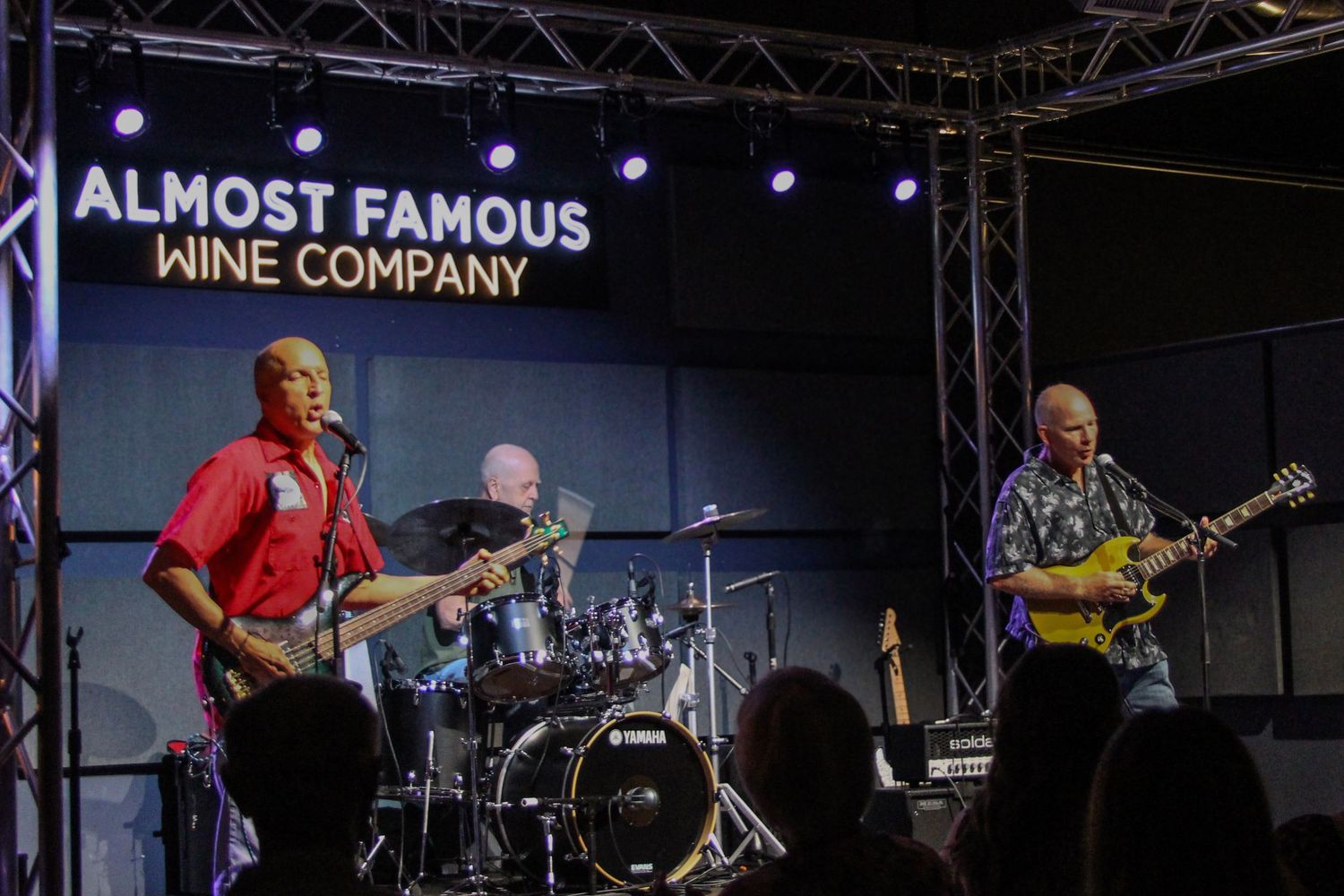 Live band performing on stage at Almost Famous Wine Company, featuring guitarists and drummer under bright lights.
