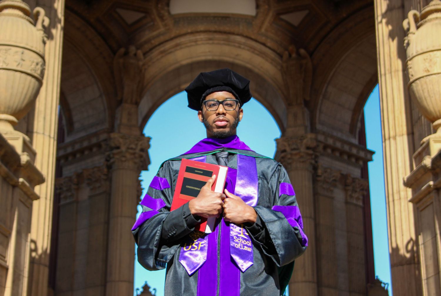 Graduate in black and purple academic regalia holds books while posing at ornate stone archway during graduation ceremony.