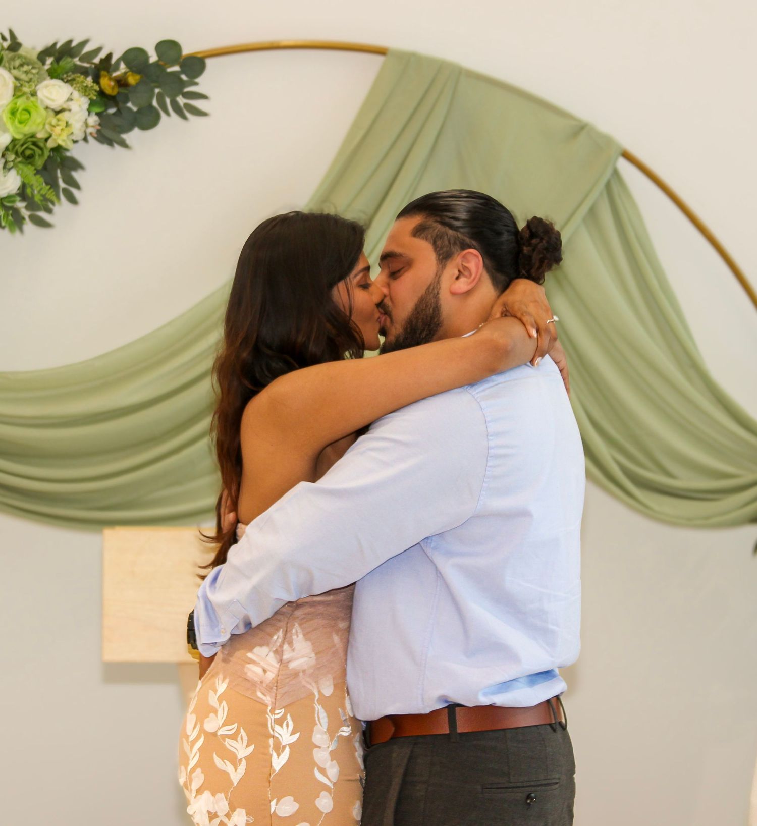Couple sharing first kiss at wedding ceremony under floral arch with sage green draped backdrop.
