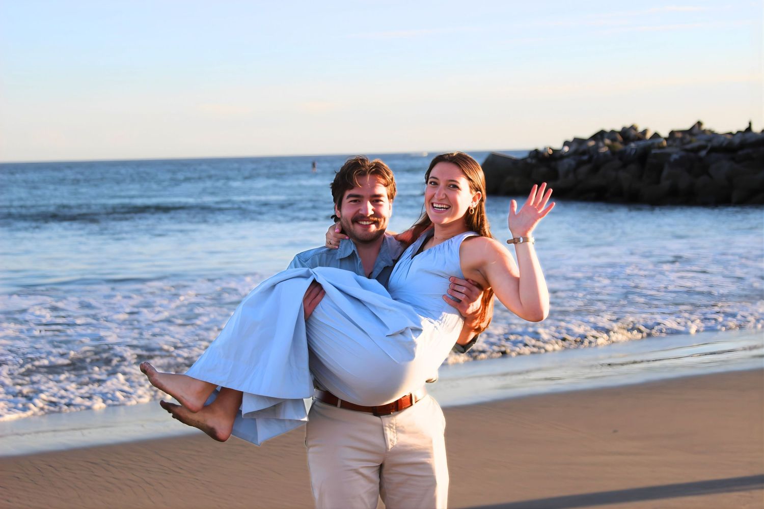 A happy couple poses on a sunny beach, man carrying woman in his arms as she waves and smiles at camera.