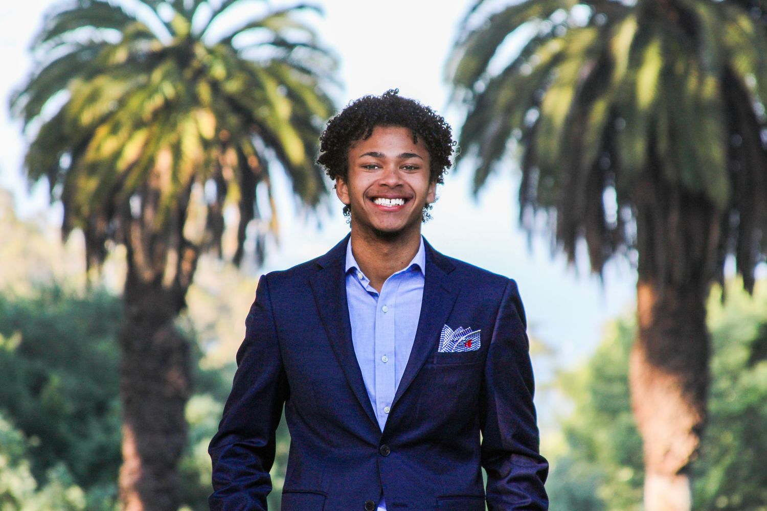Young Black man in navy suit smiling outdoors with palm trees in background on sunny day.