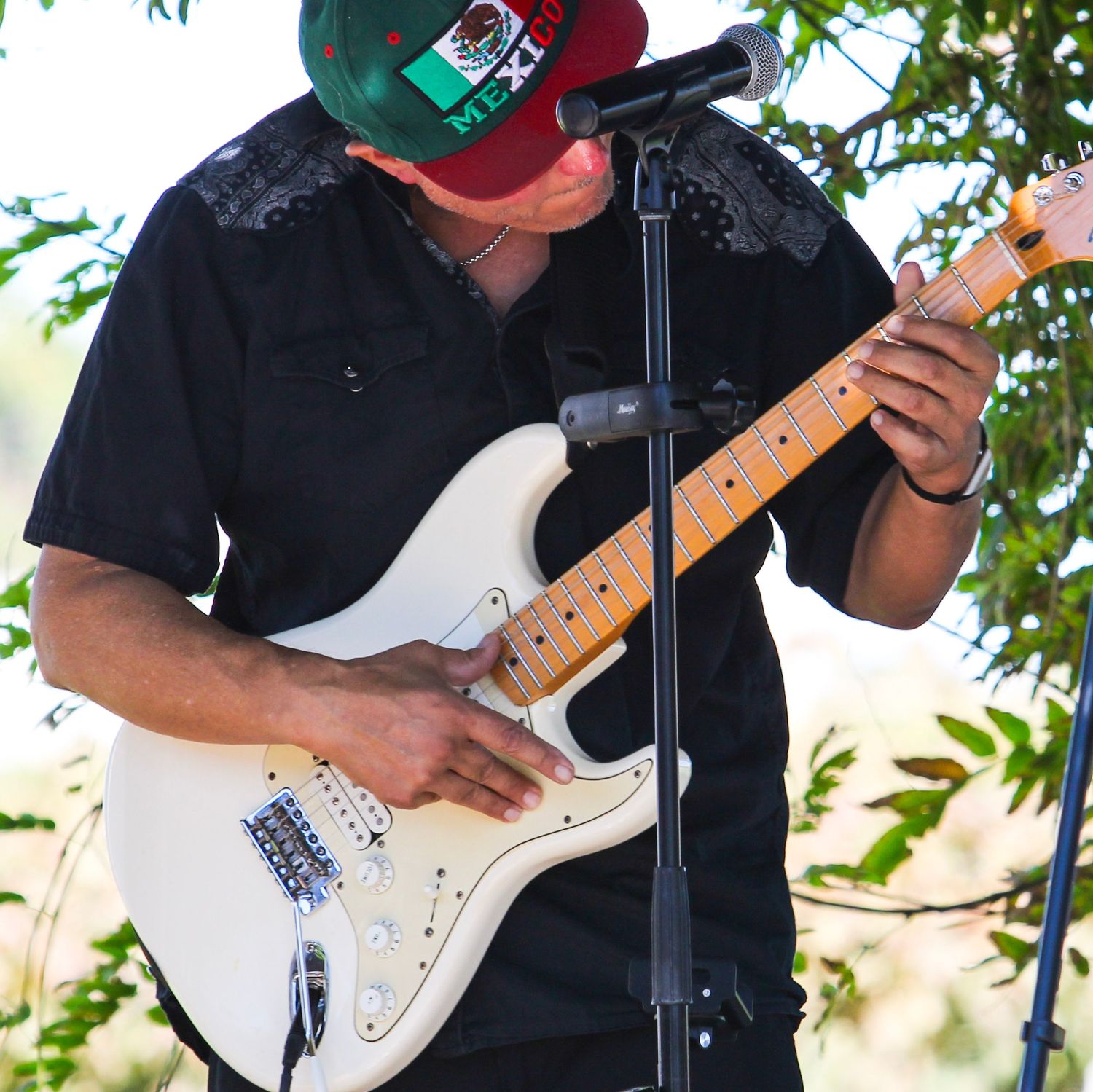 Man in black shirt and Mexico cap plays white electric guitar at outdoor microphone stand.