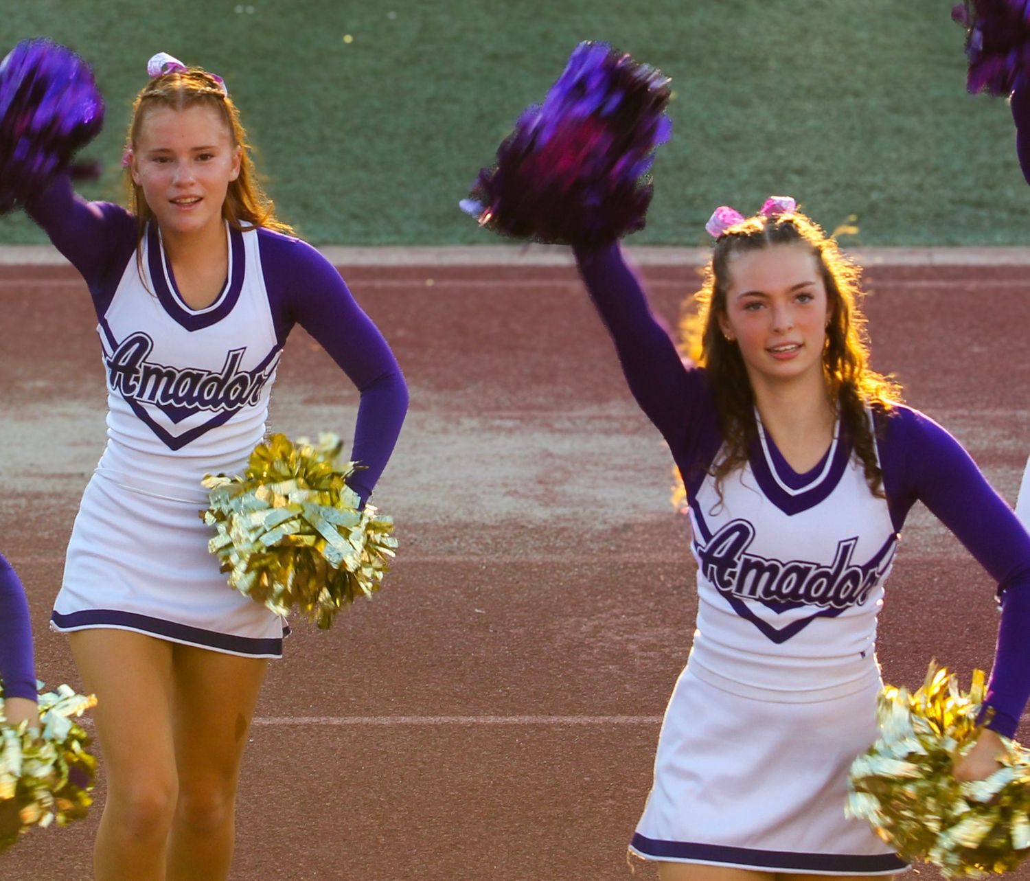 Two Amador cheerleaders in white and purple uniforms wave gold pom-poms on a track and field sideline.