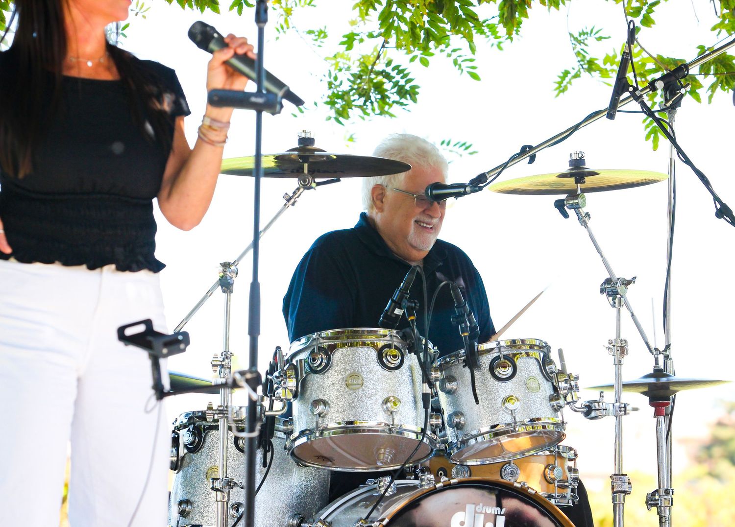 Male drummer performs on stage outdoors while female vocalist sings into microphone at live music event.