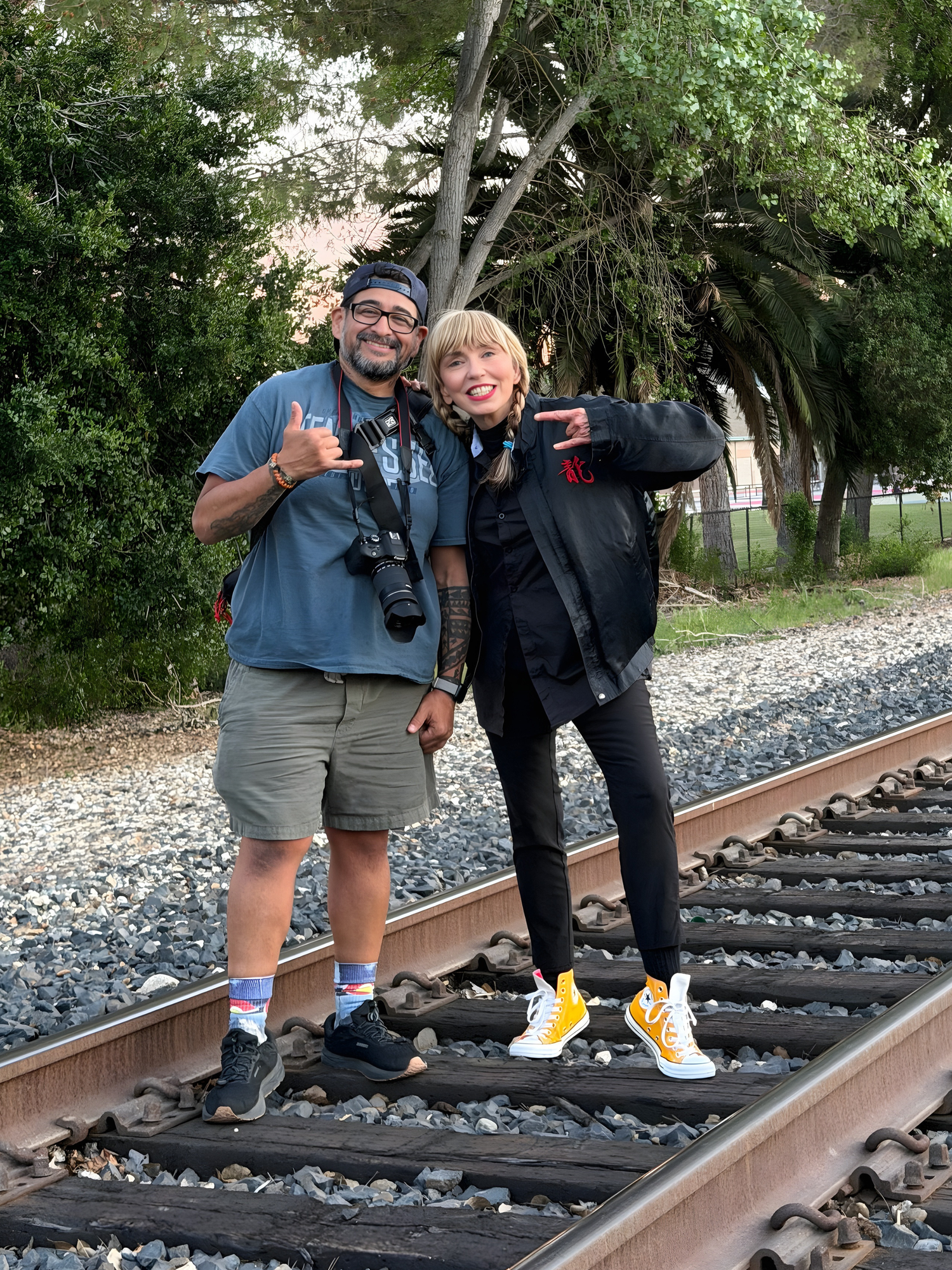 Two friends pose on railroad tracks surrounded by trees, one holding a camera, both making hand gestures.