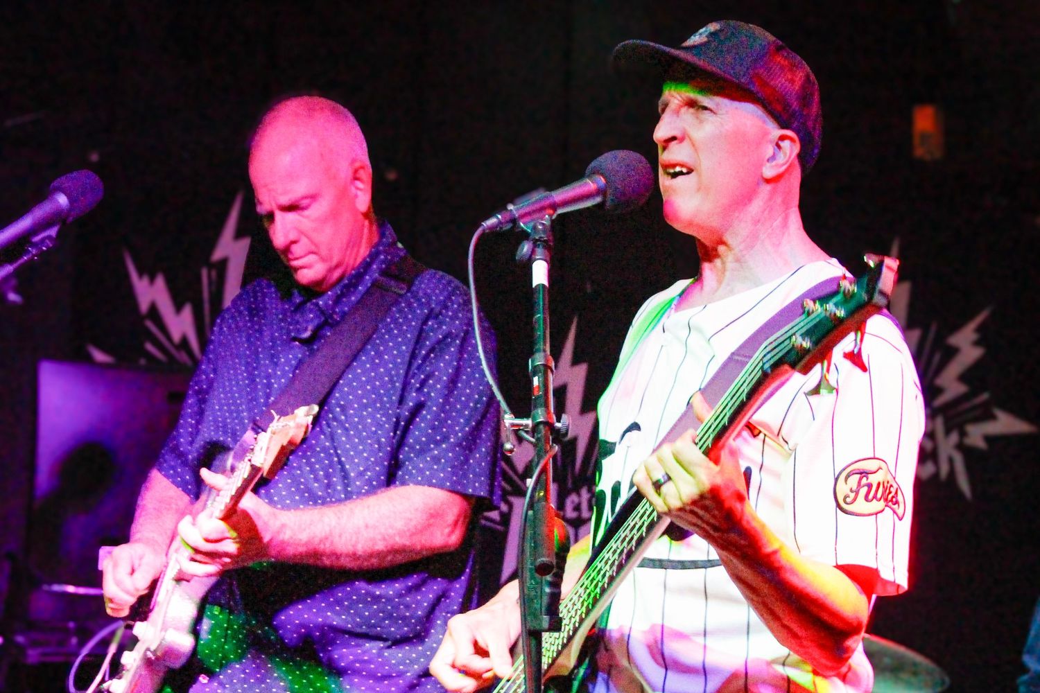 Two musicians performing on stage, one playing electric guitar in a baseball jersey, the other in a blue shirt at a live concert.