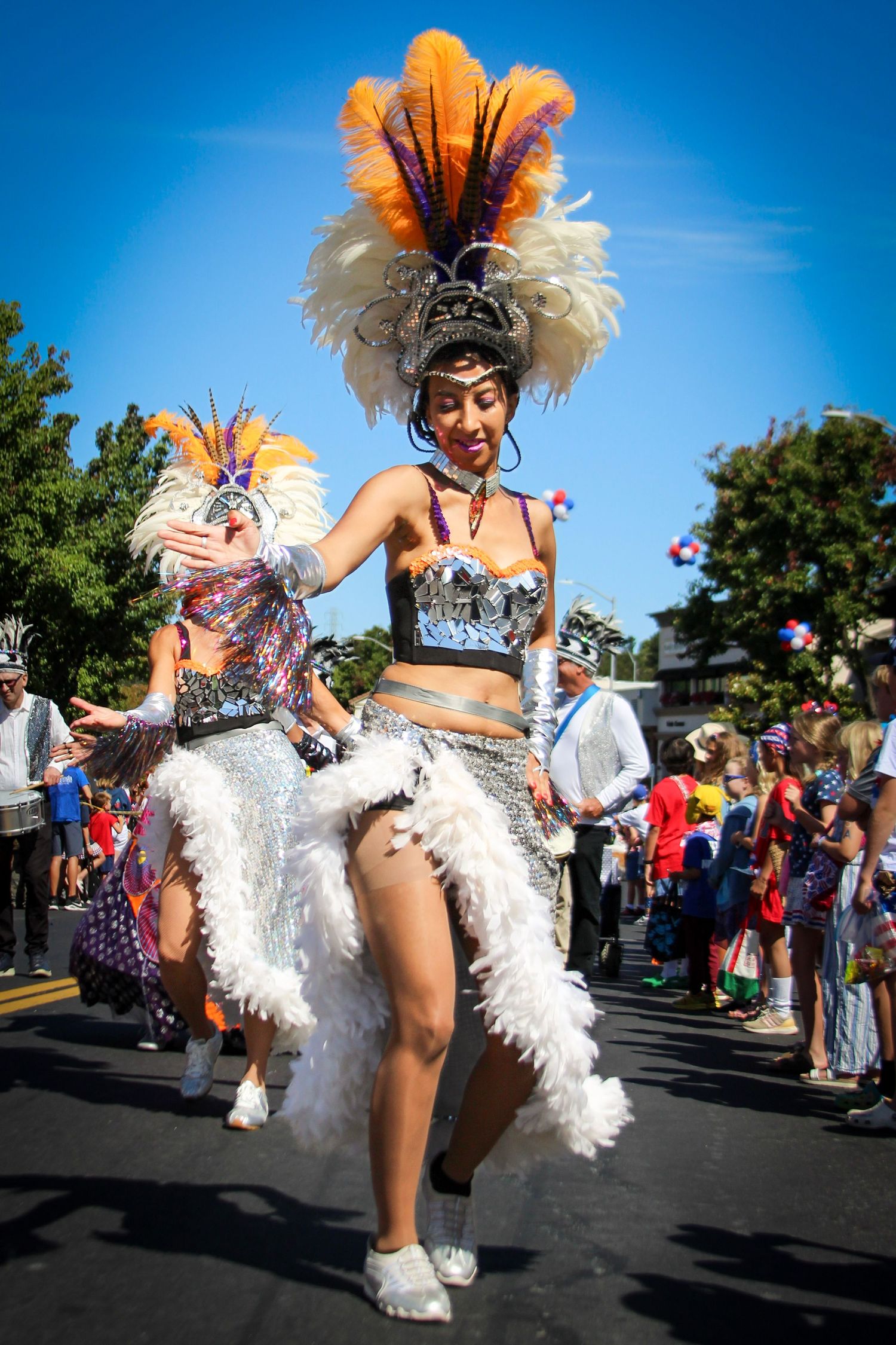 Woman in feathered carnival costume performs in colorful street parade under blue sky.