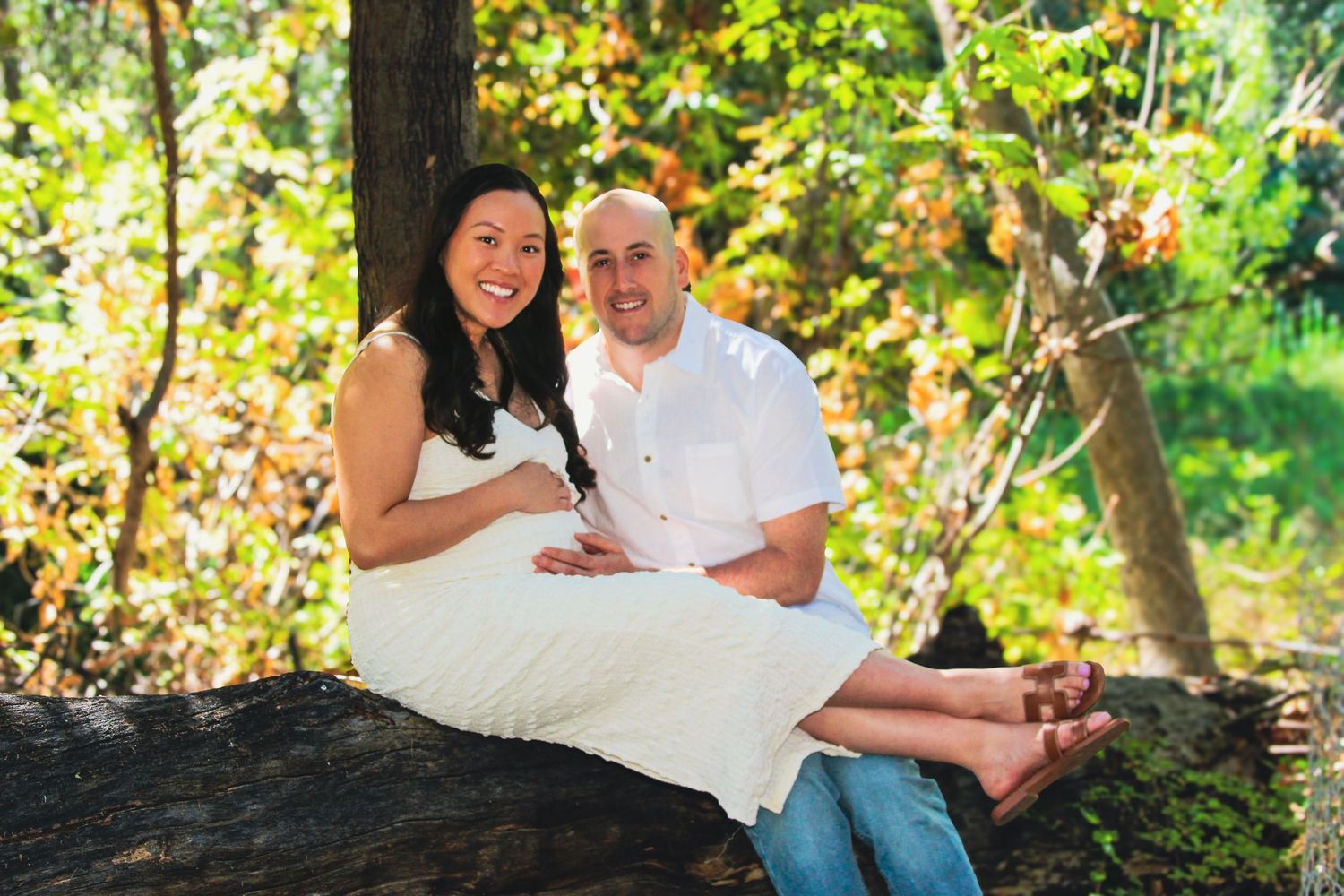 Happy couple sitting on a tree log outdoors, surrounded by colorful autumn foliage in a natural park setting.