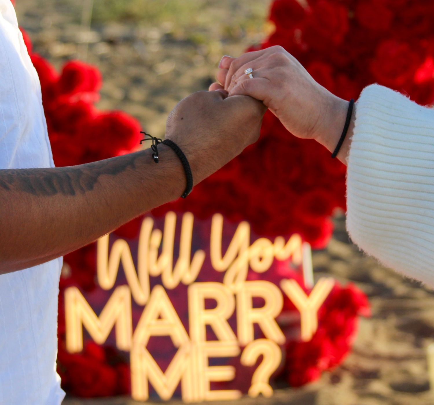 A romantic marriage proposal scene with illuminated Will You Marry Me sign surrounded by red roses as a couple holds hands.