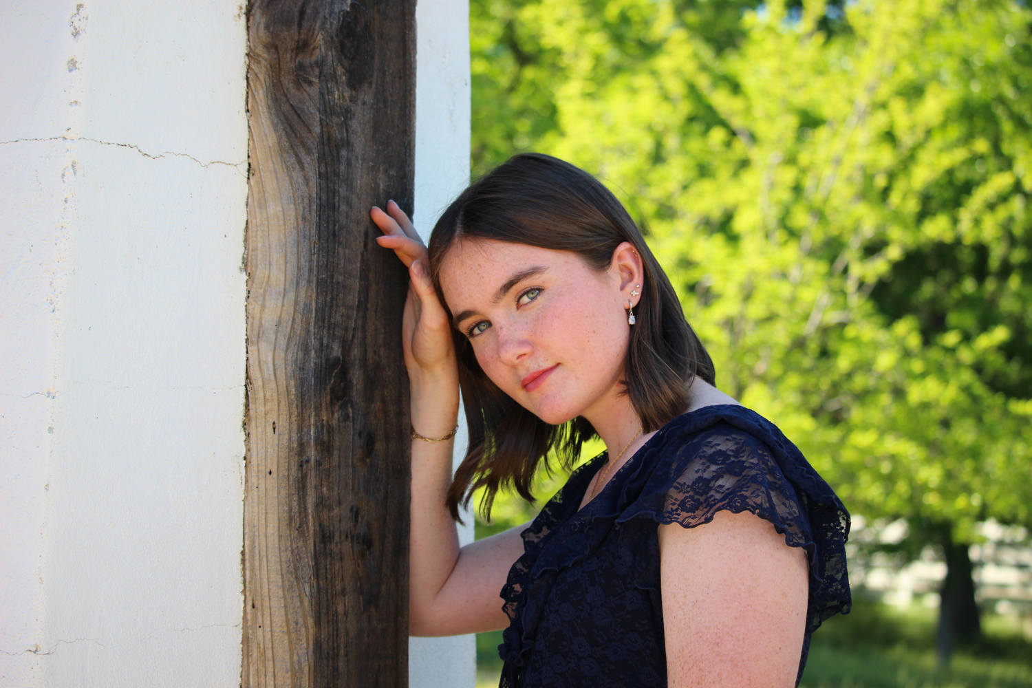 A young brunette woman in a navy lace dress leans against a wooden post outdoors in soft sunlight. portraits, photographer