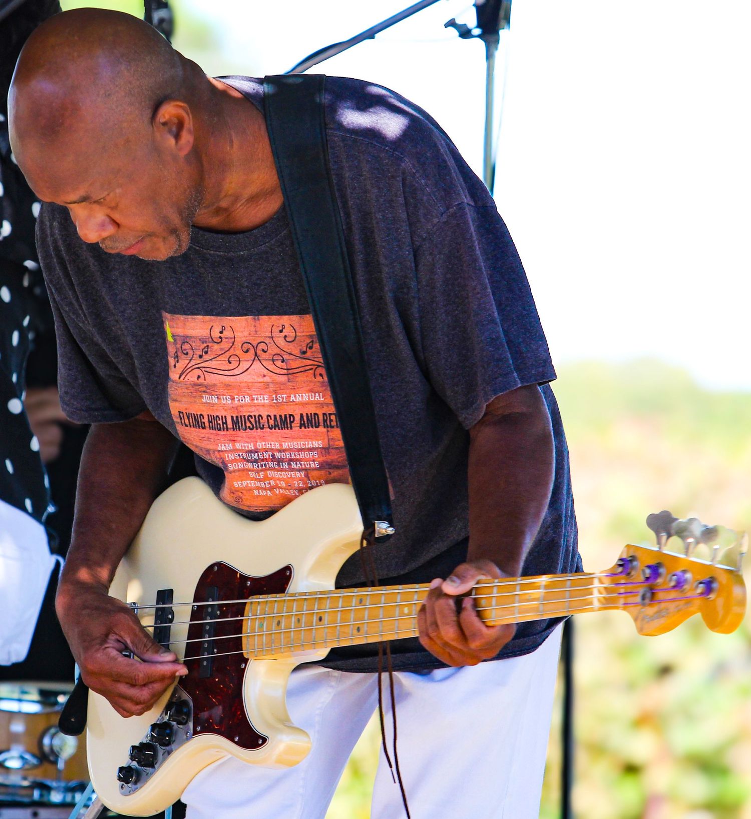 Man playing a cream-colored bass guitar on stage outdoors, wearing a gray t-shirt, focused on his performance.