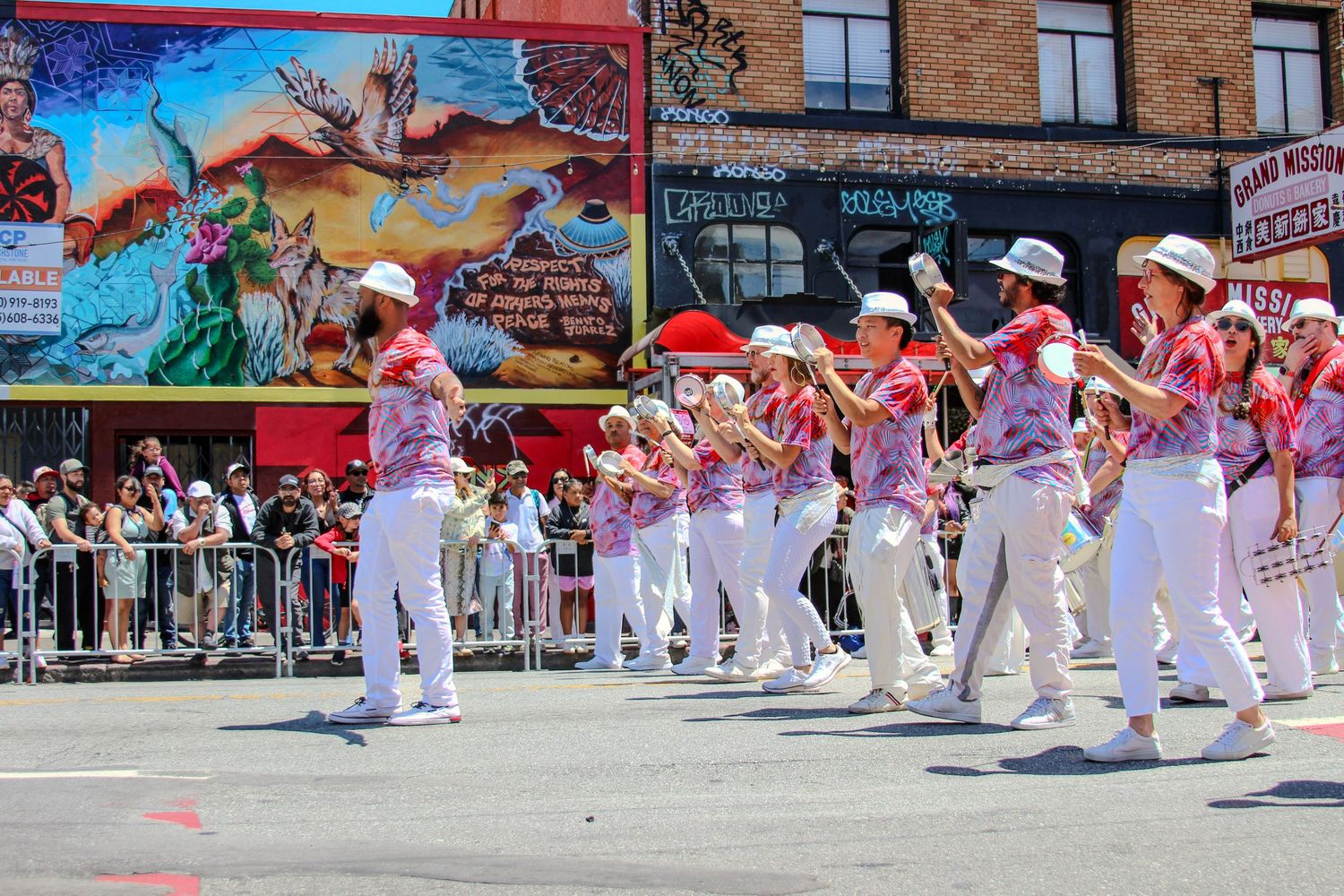 Marching band in white and pink uniforms performing in a colorful street parade with festive murals in the background.