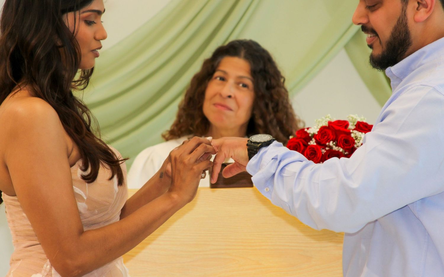 Couple exchanging rings during an intimate wedding ceremony officiated by a smiling woman in a green-draped venue.