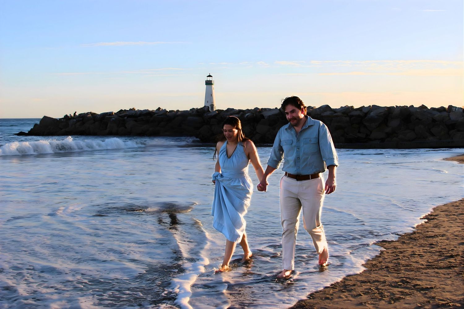 A couple walks hand in hand along a sandy beach at sunset with a lighthouse visible behind them. santa cruz, surprise engagement, photograph