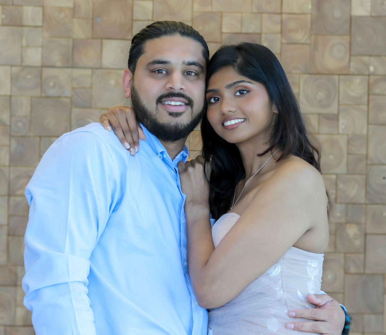 Smiling couple embracing closely in front of a wooden tile wall, woman in pink dress, man in blue shirt.