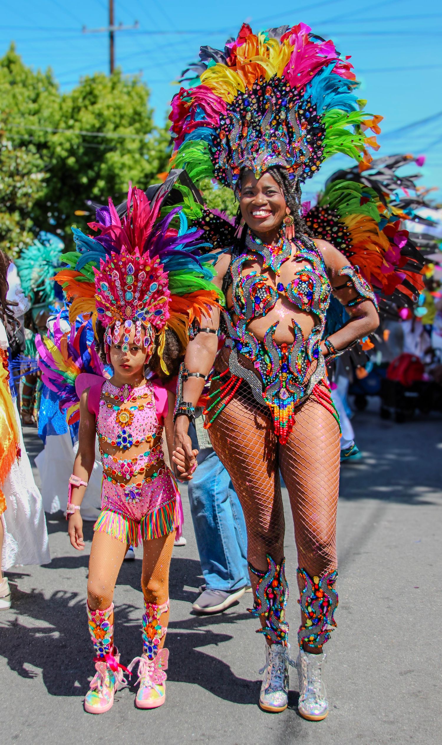 Woman and girl in vibrant rainbow feathered carnival costumes parade together at a colorful street festival.