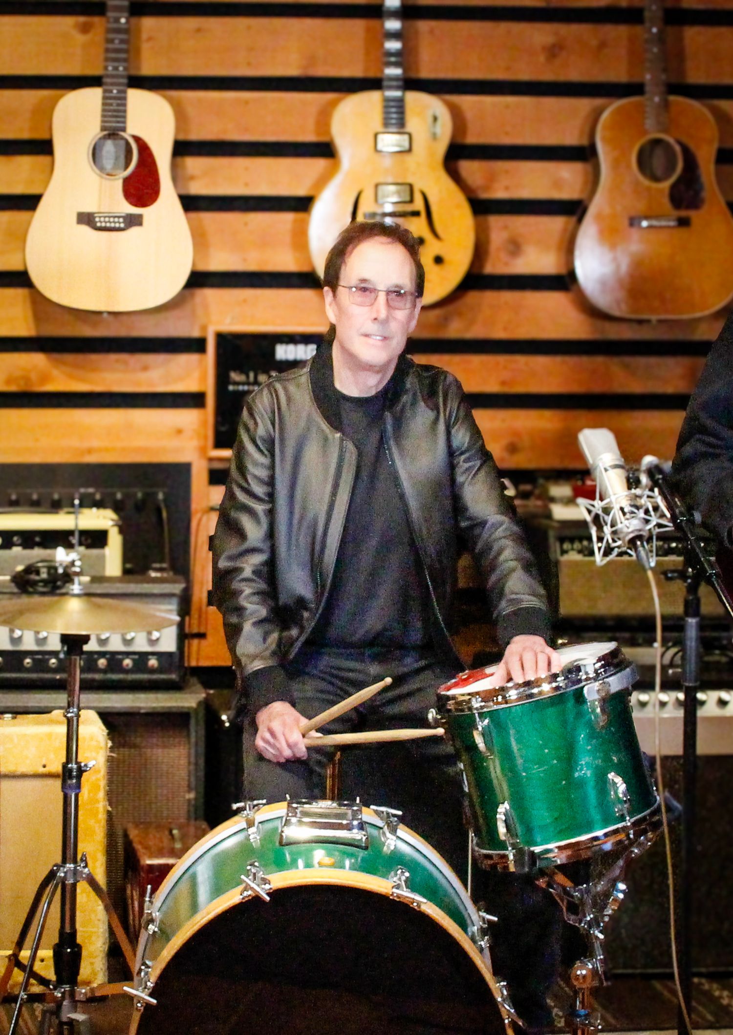 Musician playing green drum kit in a studio surrounded by acoustic and electric guitars on wooden wall.