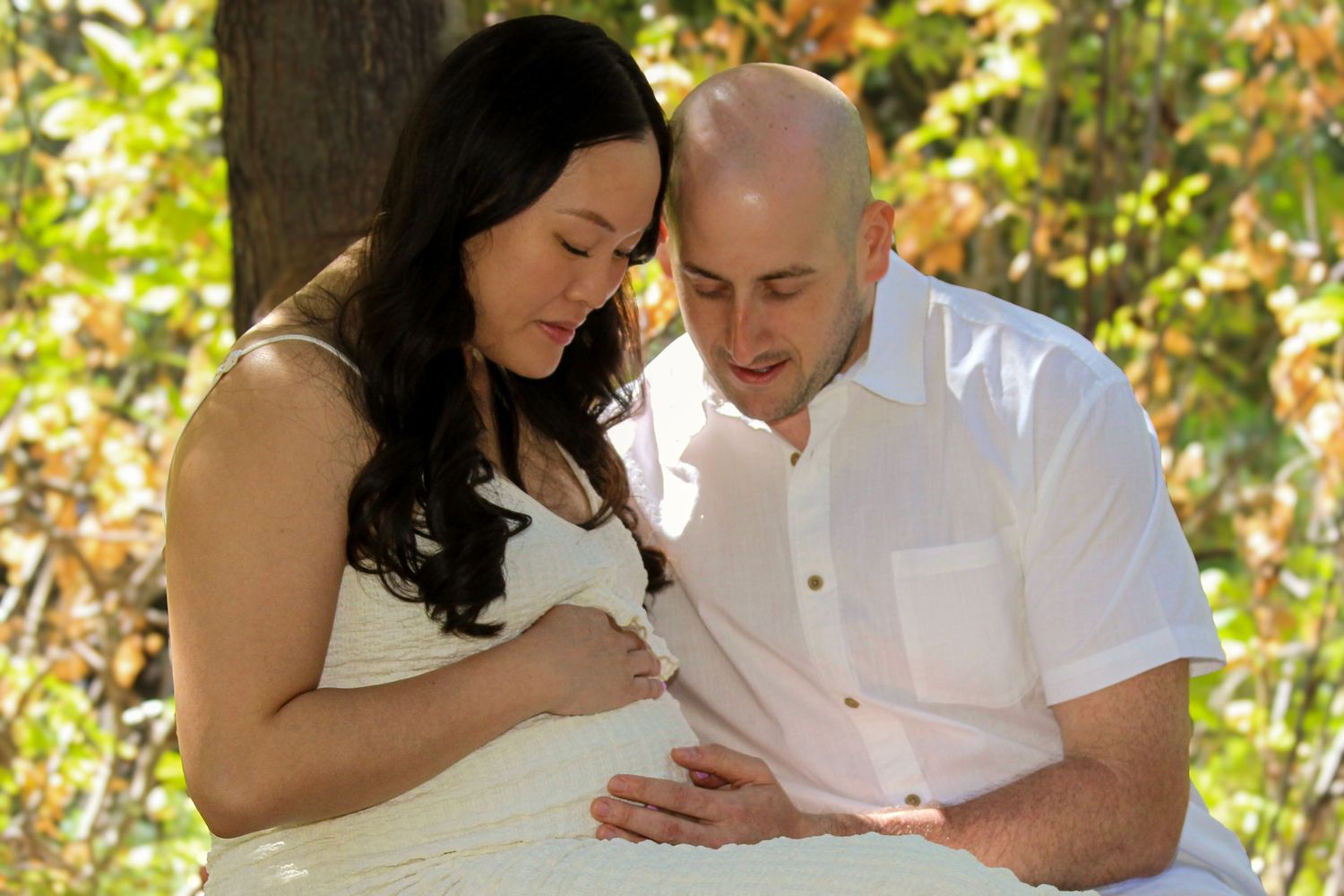 Expectant couple in white outfits sharing a tender moment outdoors, looking at pregnant belly among autumn foliage.