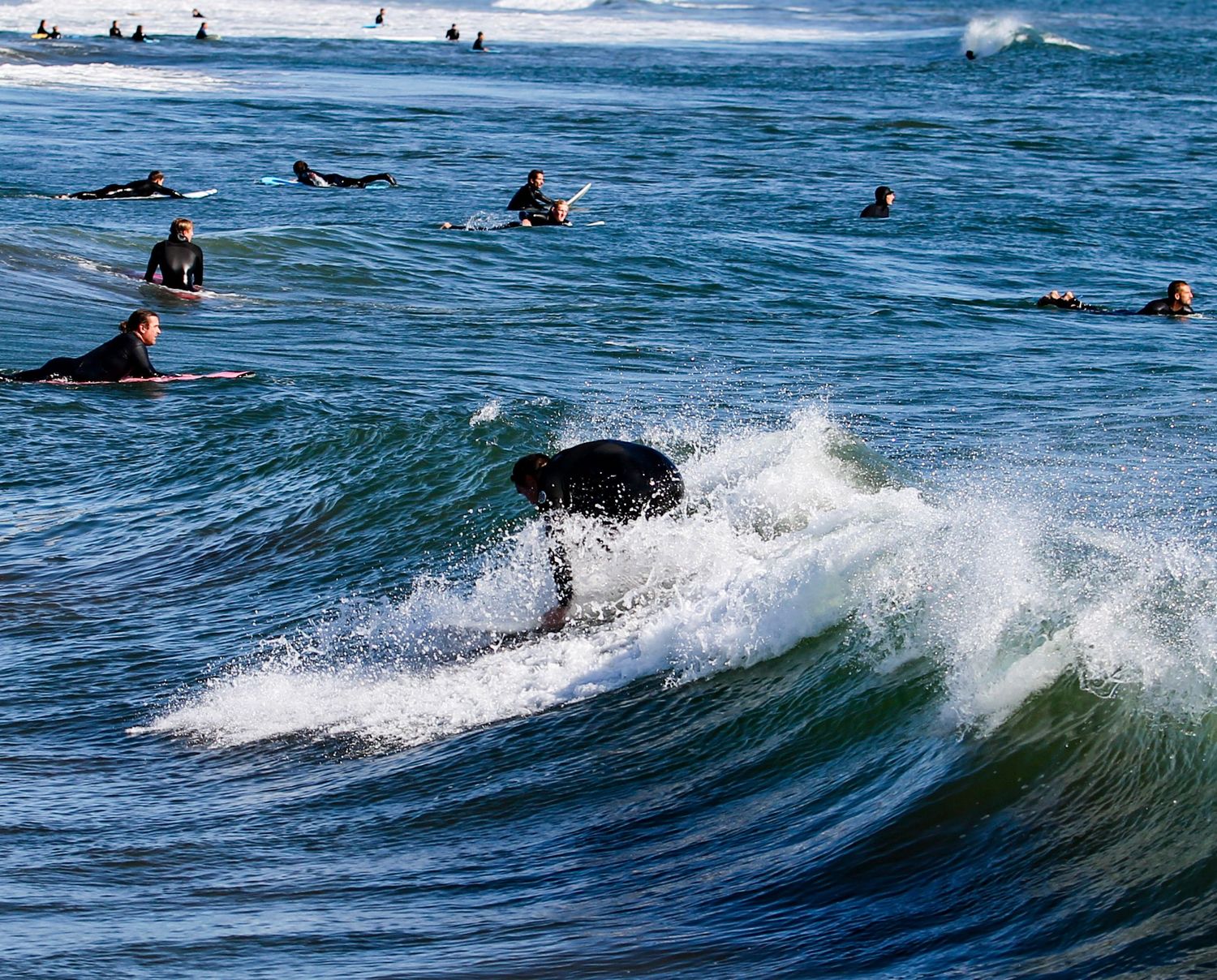 Surfers in wetsuits riding waves and paddling in the ocean at a busy California surf break.