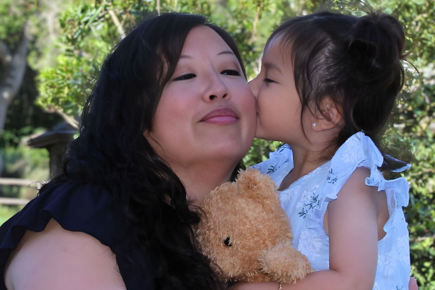 A woman holds a toddler girl with a teddy bear as the child gives her a sweet kiss on the cheek. lake chabot, mommy and me. east bay