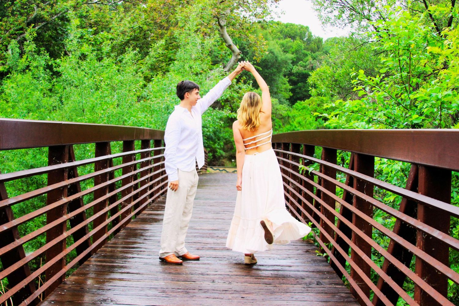 Couple dancing on a wooden bridge surrounded by lush green trees in a romantic outdoor setting.