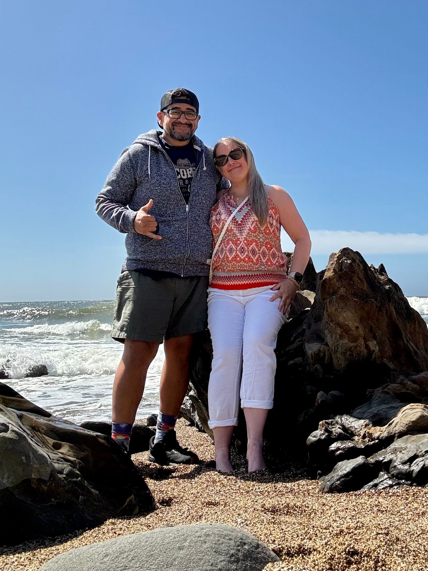 Couple posing on rocky beach with ocean waves, man in hoodie giving thumbs up, woman in white capris and colorful top.