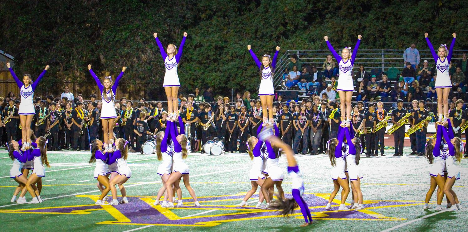 Cheerleaders perform pyramid stunts in purple and white uniforms on a football field during a nighttime pep rally.