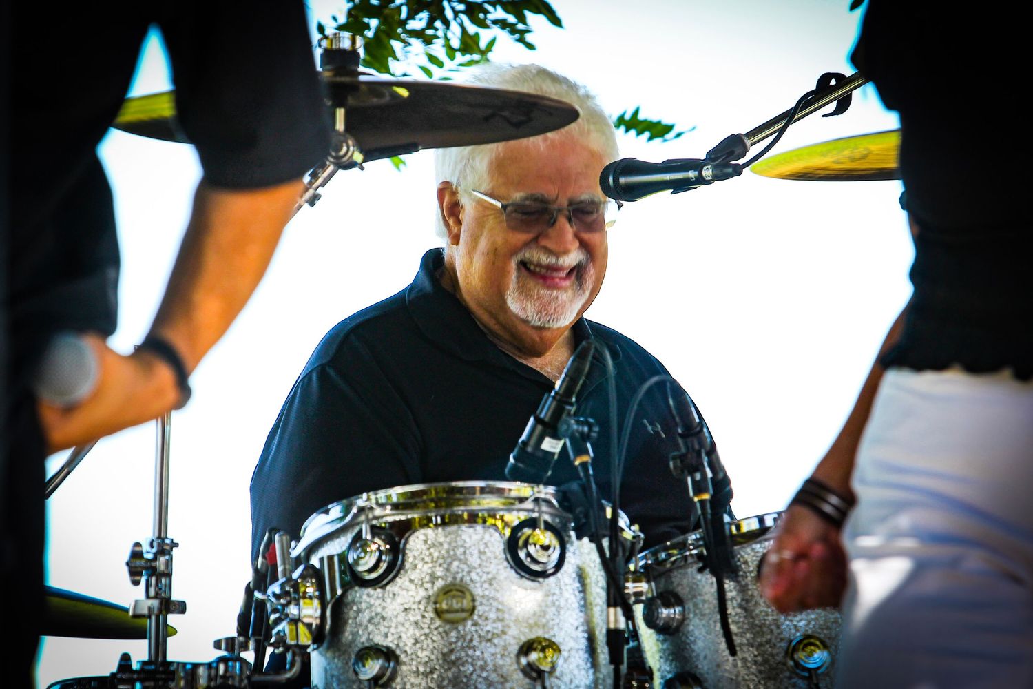 Smiling senior man playing drums at an outdoor music event, wearing glasses and a black shirt.