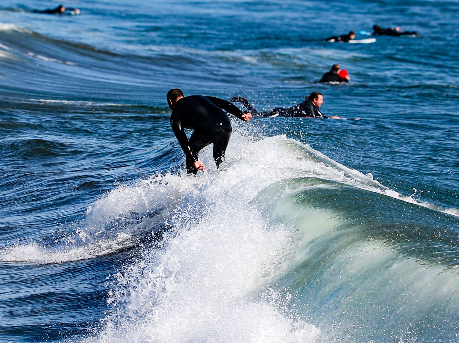 Surfer in black wetsuit riding ocean wave with other surfers paddling in background at sunny beach.