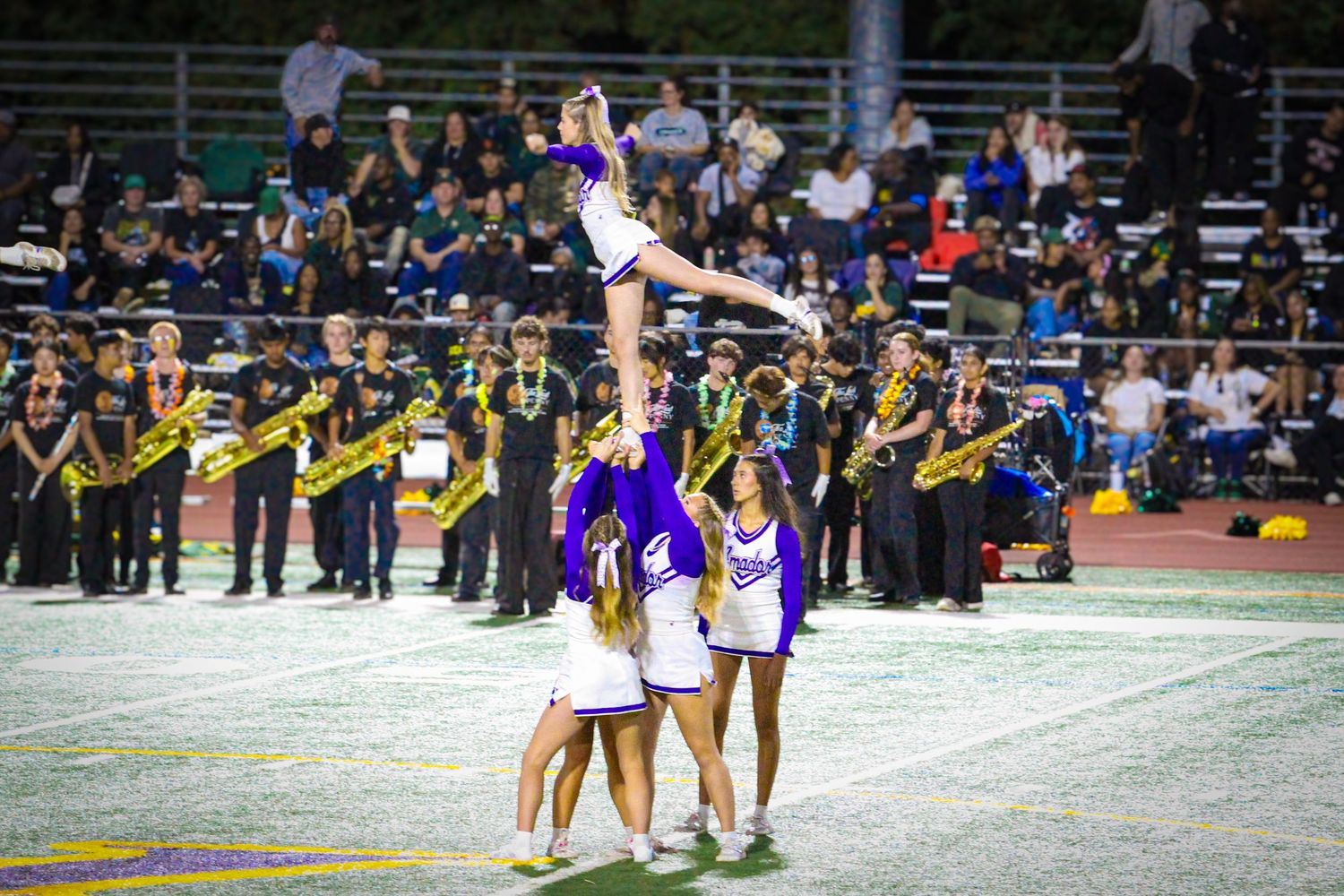Cheerleaders perform a stunt lift at a night football game with a marching band in purple uniforms in the background.