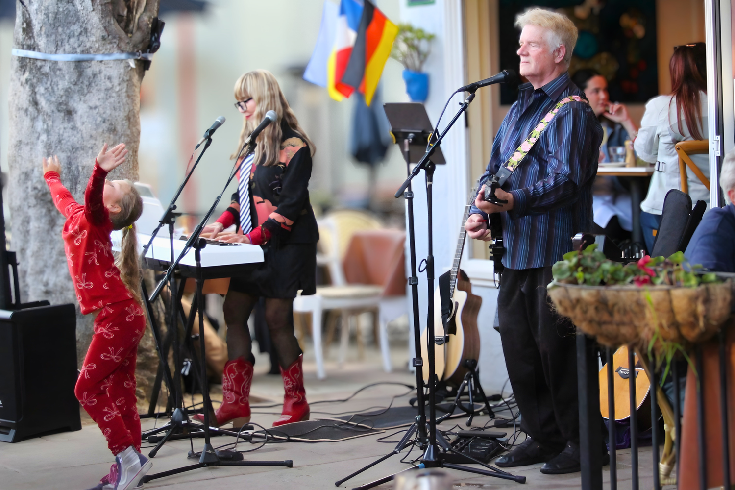 Live outdoor band performance with guitarist, female vocalist, and dancing child in red pajamas at a festive venue.
