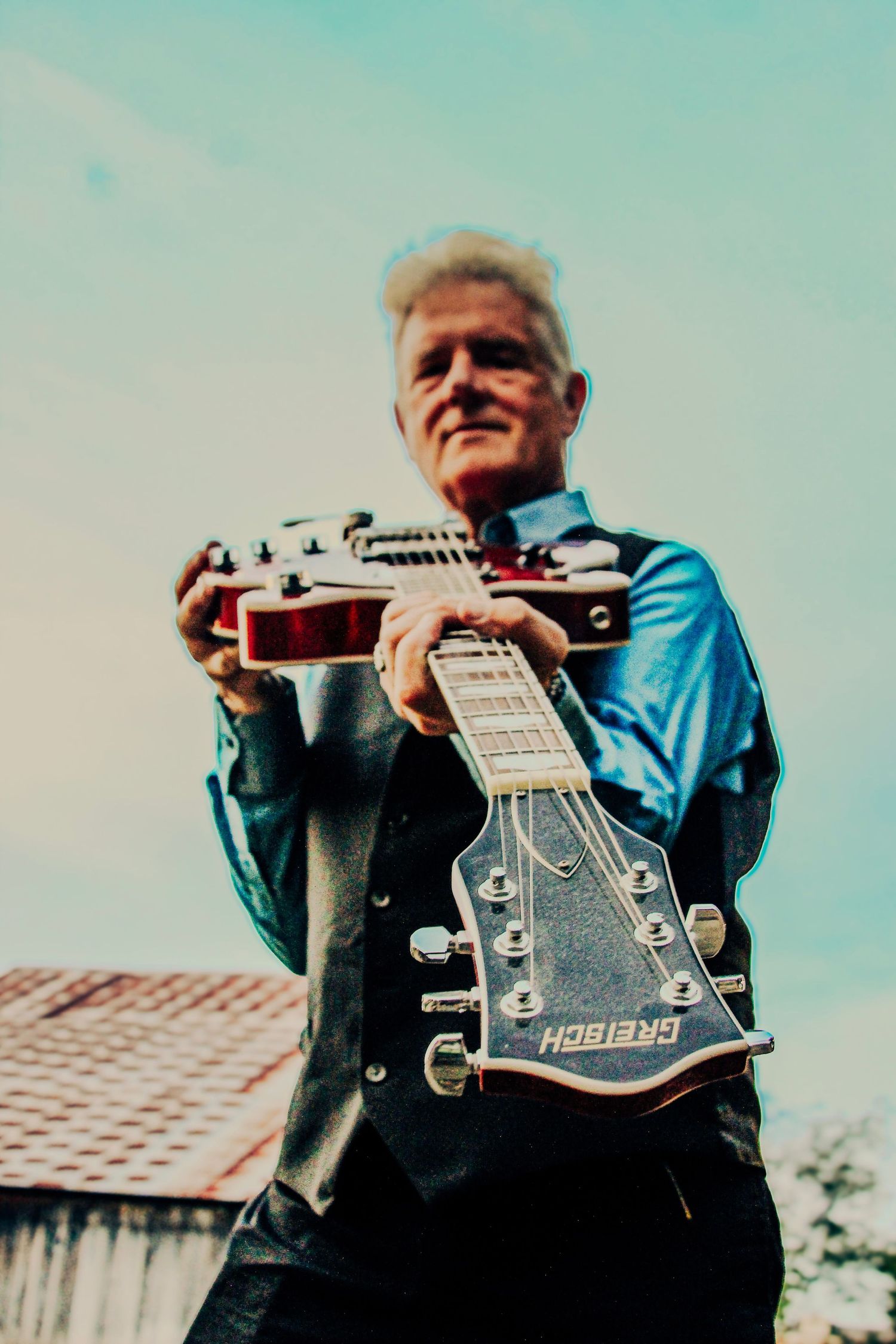 Man in blue shirt holds Gretsch electric guitar outdoors, low-angle shot against sky.