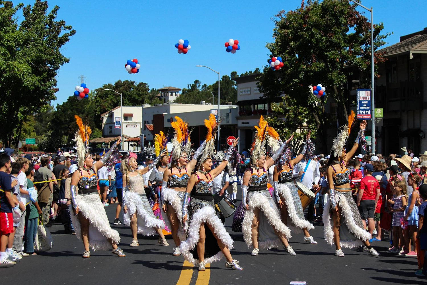 Colorful parade performers in feathered costumes dance down a sunny street with red, white, and blue balloons overhead.
