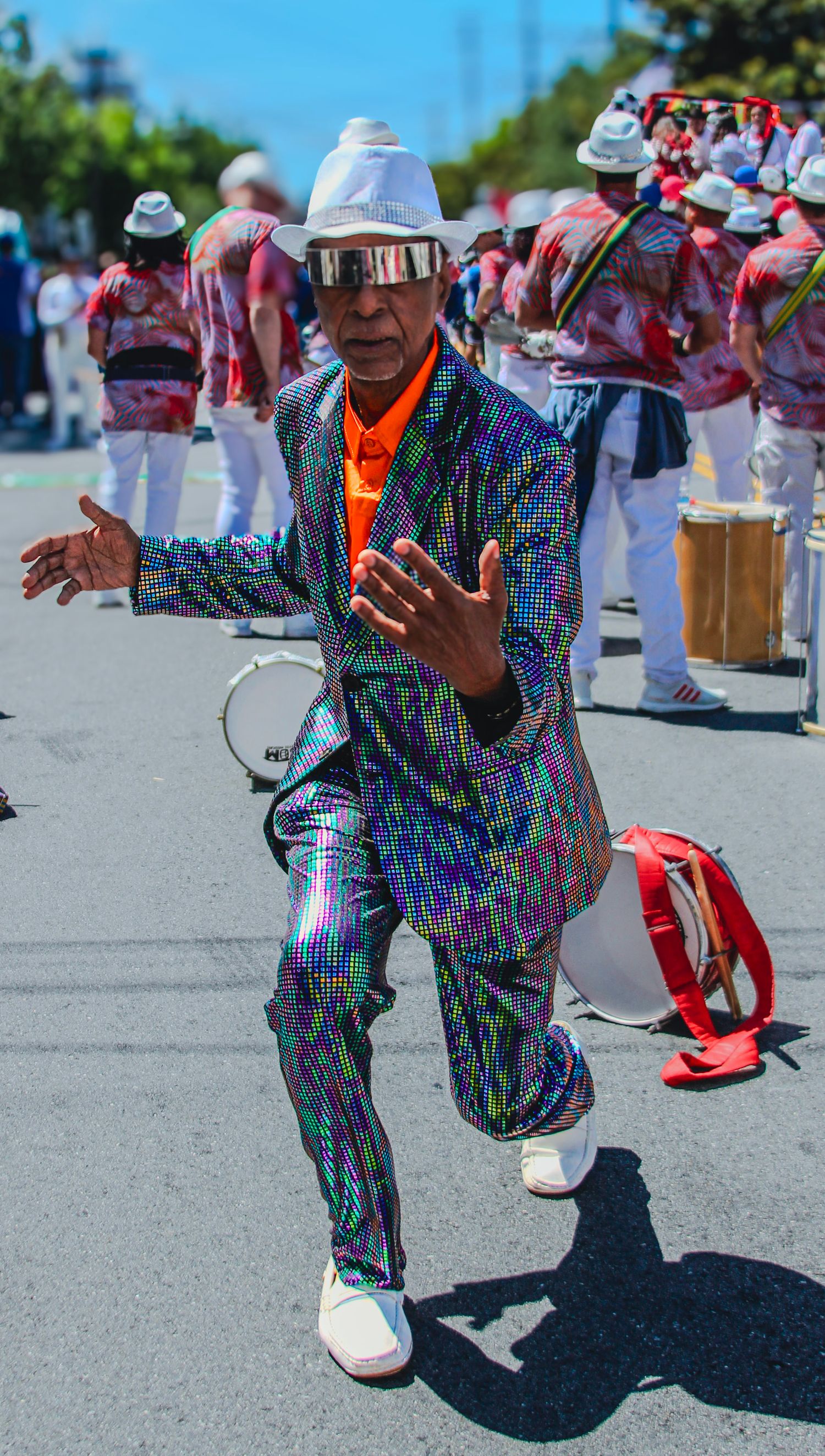 Man in iridescent sequin suit and white hat dancing at a vibrant outdoor carnival parade with drummers.