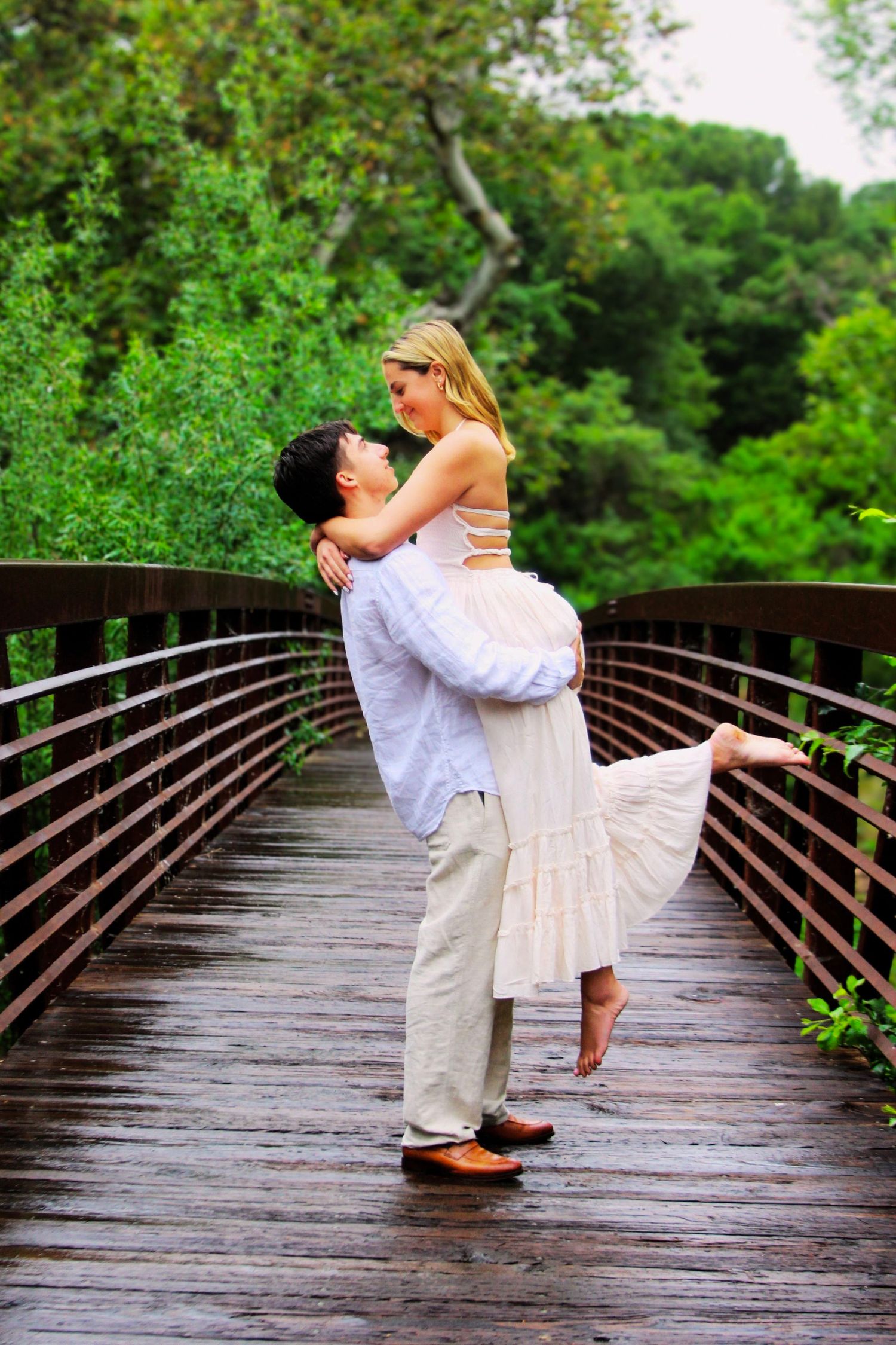 A romantic couple embraces on a wooden bridge surrounded by lush green trees, as the man lifts the woman in a white dress.