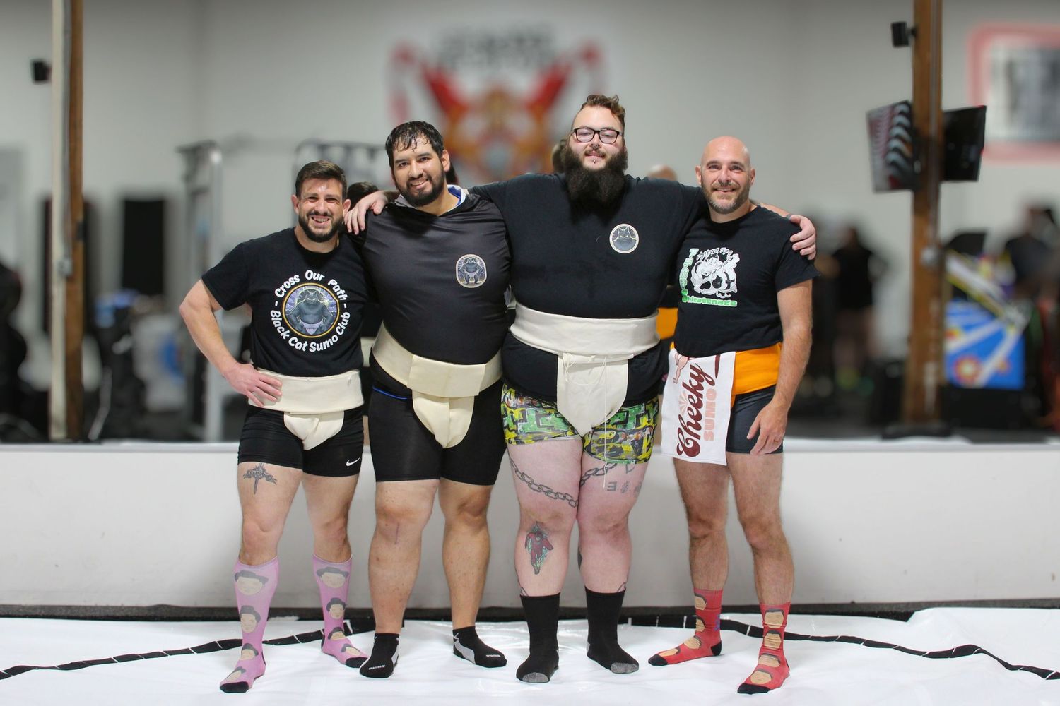 Four men in sumo wrestling costumes pose together smiling in a gym setting.