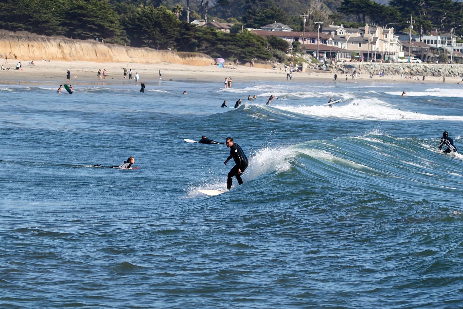 Surfer riding a wave at a busy beach with swimmers and coastal cliffs in the background.