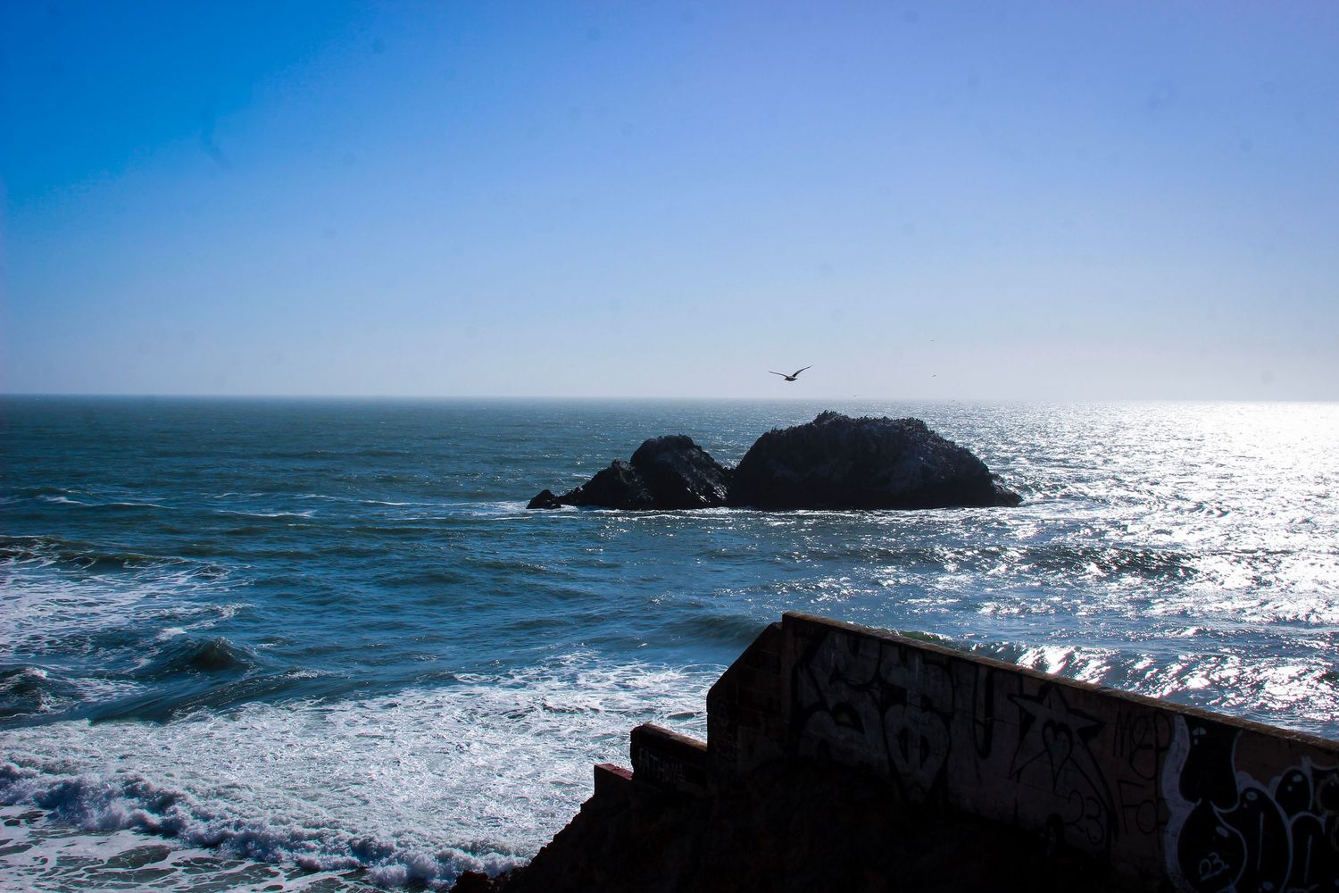 Rocky islands emerge from the Pacific Ocean waves near a graffiti-covered seawall under a clear blue sky.
