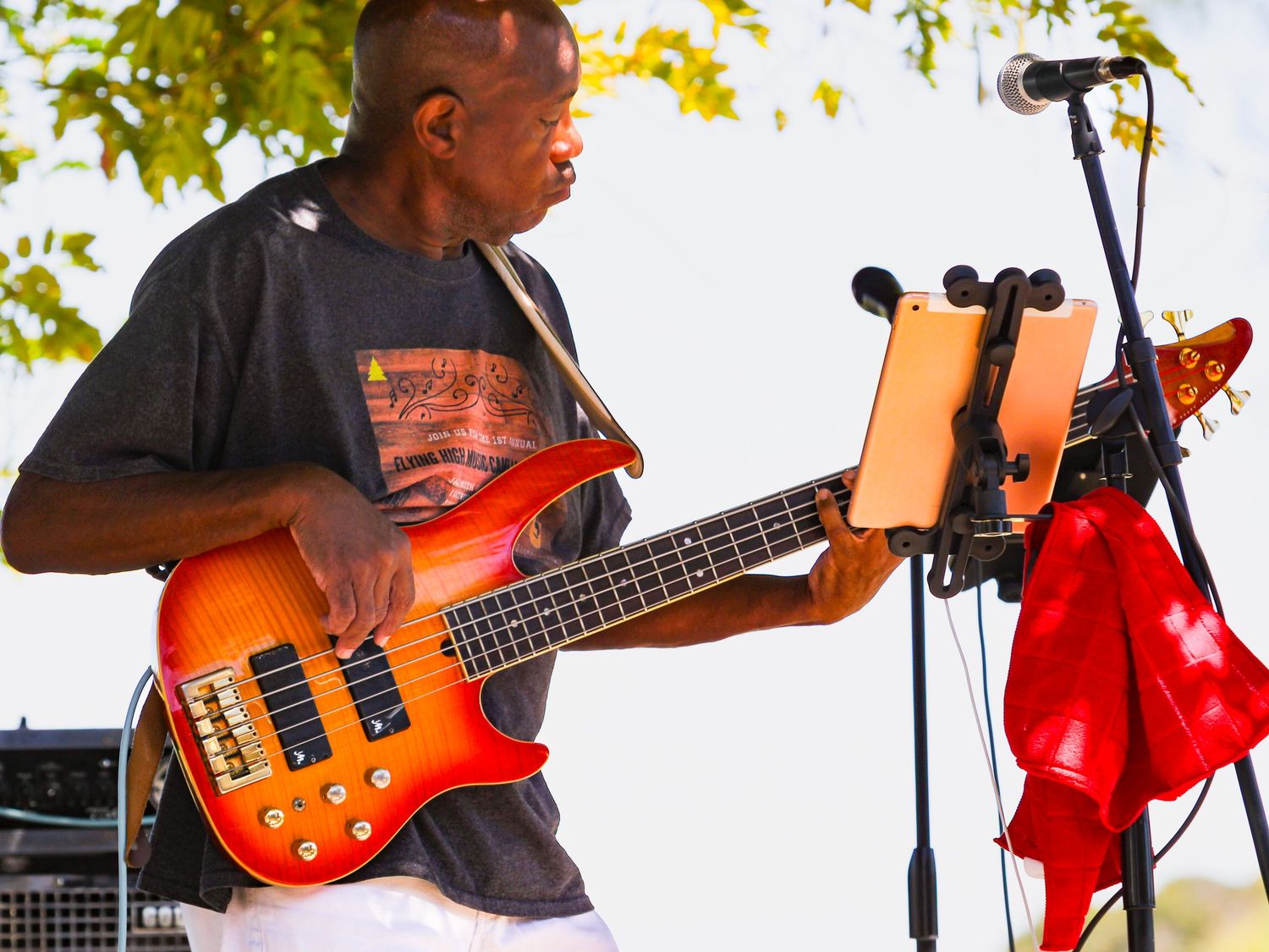 A musician plays a red electric bass guitar on an outdoor stage with a tablet on a music stand nearby.