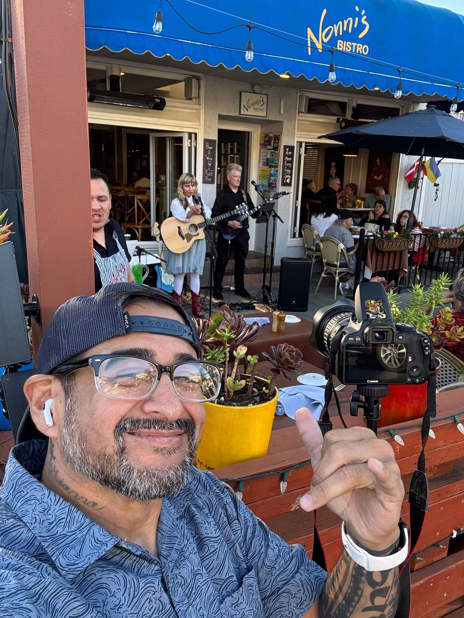 Man taking selfie at Nonni's Bistro patio with live musicians performing guitar in background.