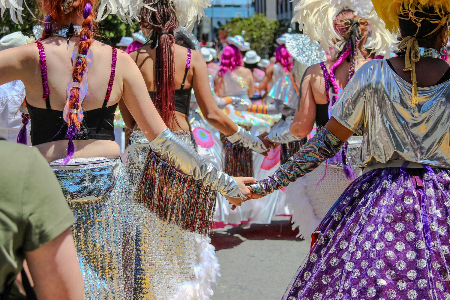Colorful carnival dancers in festive costumes with fringe and feathers hold hands during a vibrant street parade celebration.
