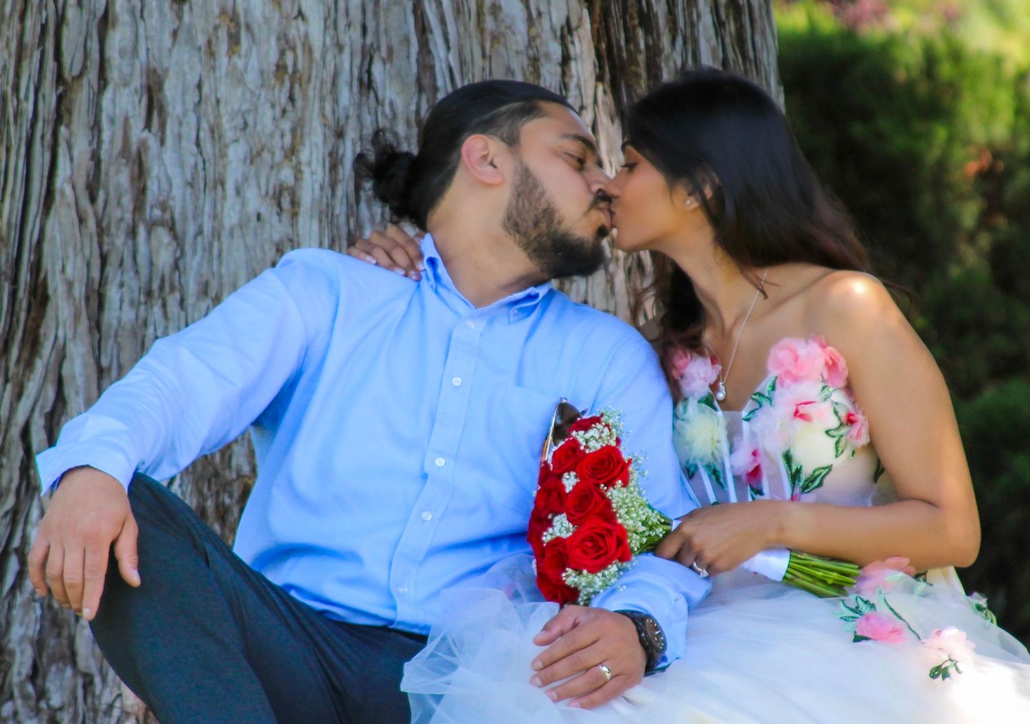 Couple sharing romantic kiss by tree, bride in floral gown holding red bouquet on wedding day.