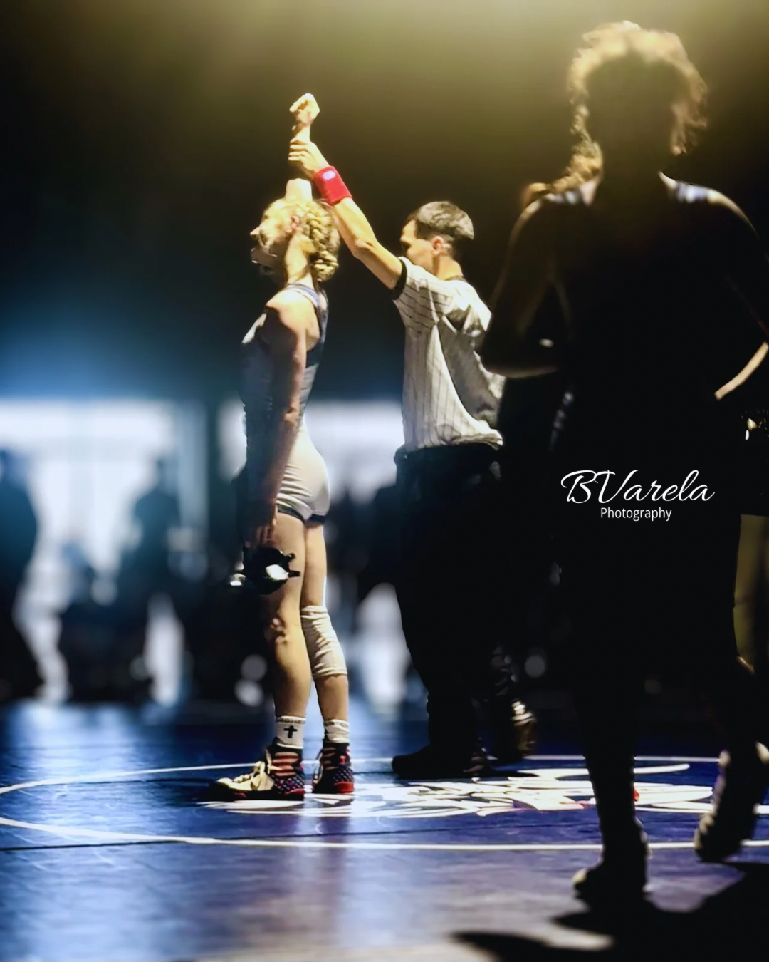 Wrestler raises opponent's arm in victory on mat under dramatic stage lighting at a wrestling tournament.