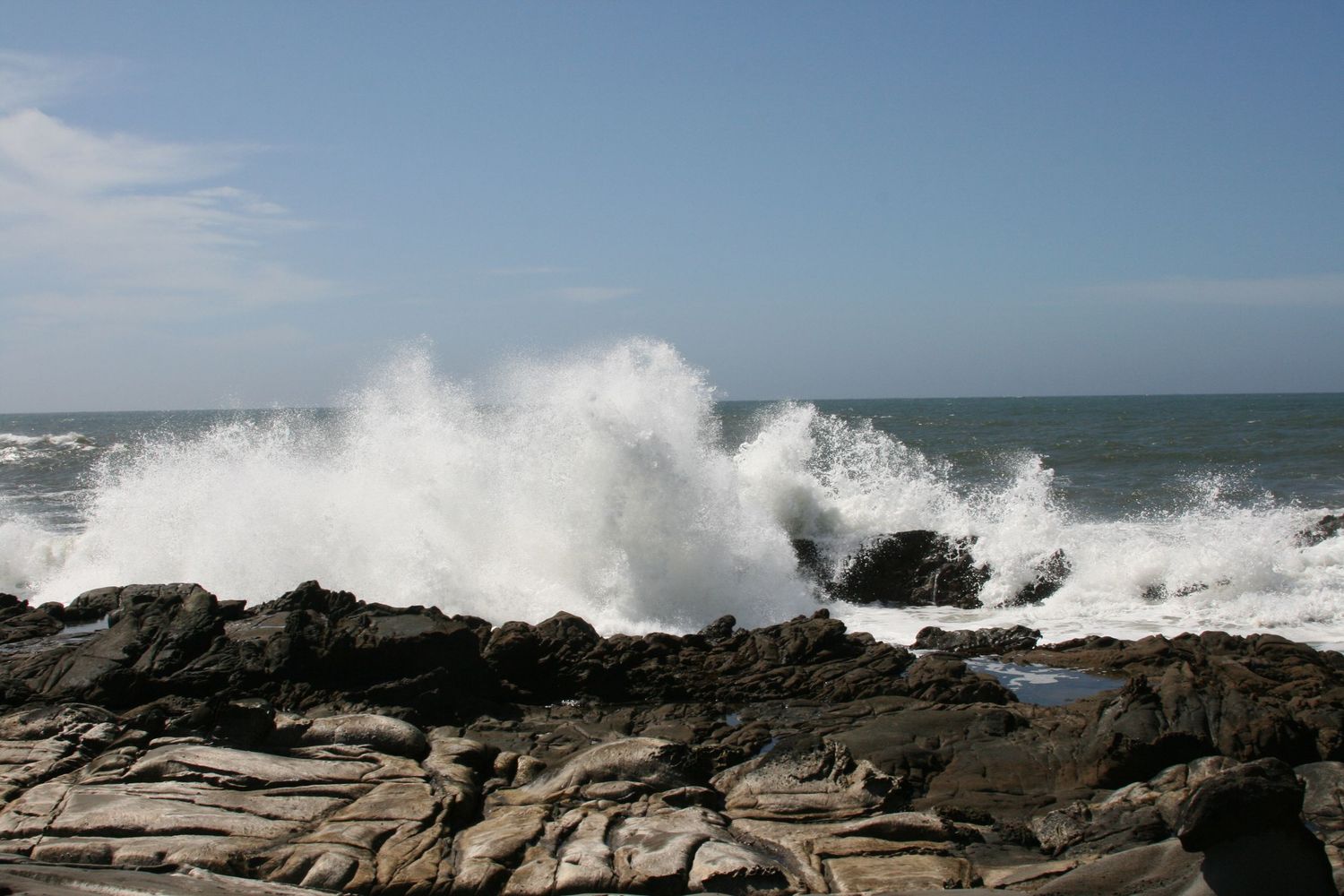 Ocean waves crashing dramatically against rocky shoreline with white sea spray under a clear blue sky.