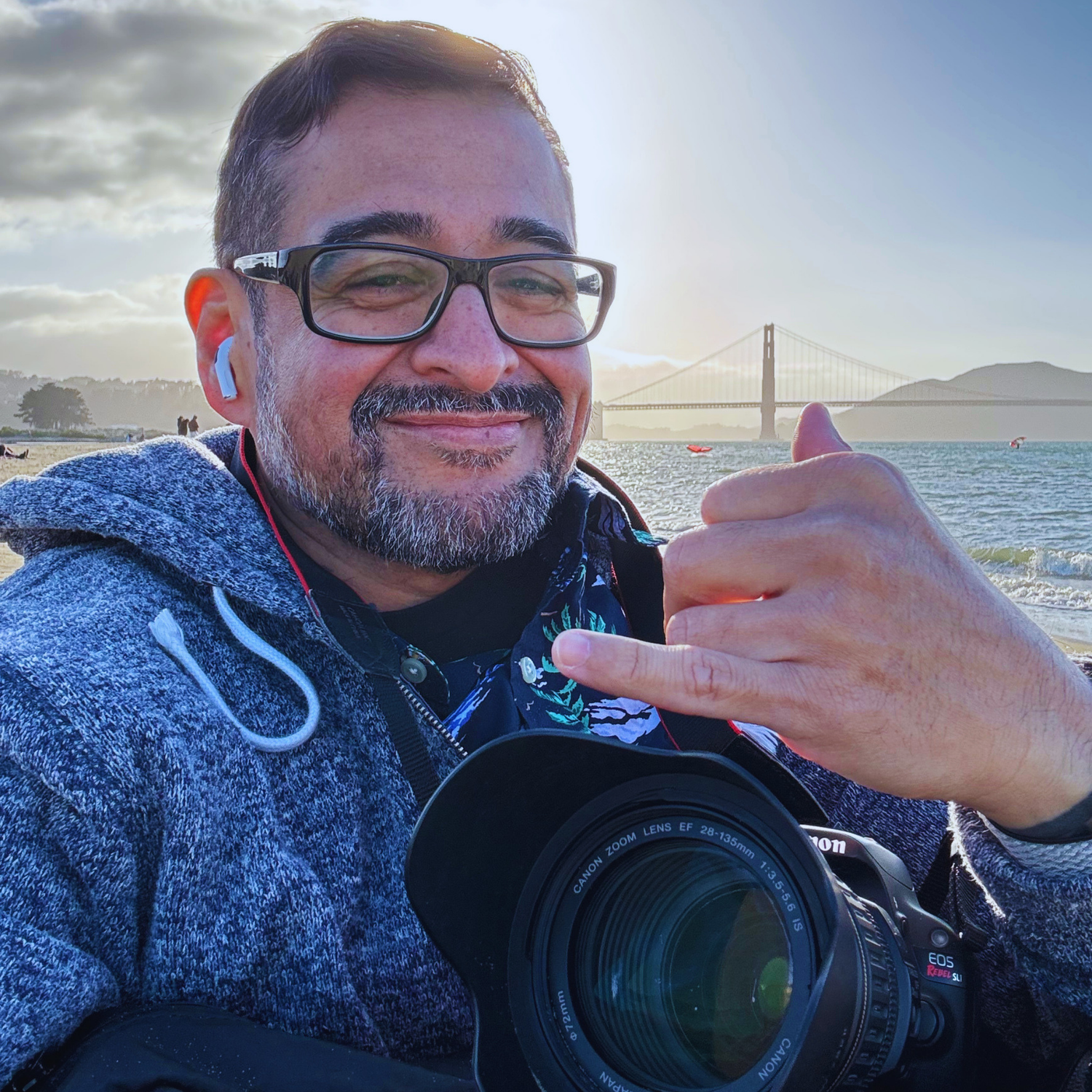 Man with glasses holding a camera gives thumbs up near the Golden Gate Bridge in San Francisco on a sunny day. tri-valley photographer