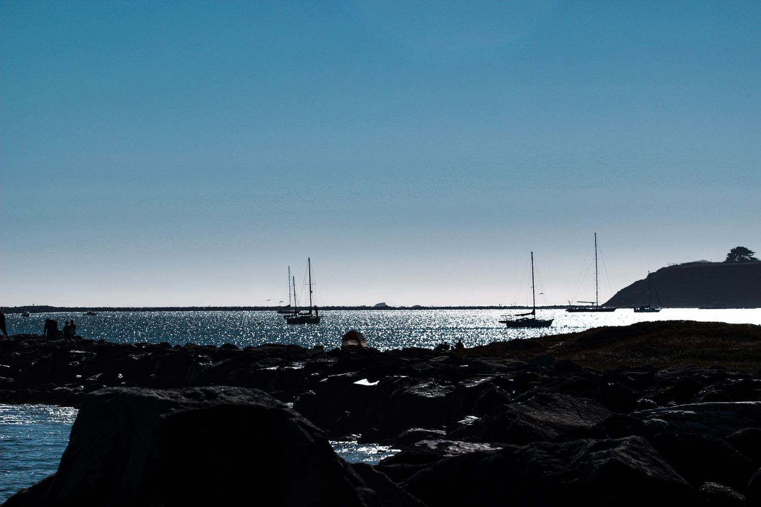Silhouetted sailboats anchored in calm blue waters near rocky coastline under clear sky.