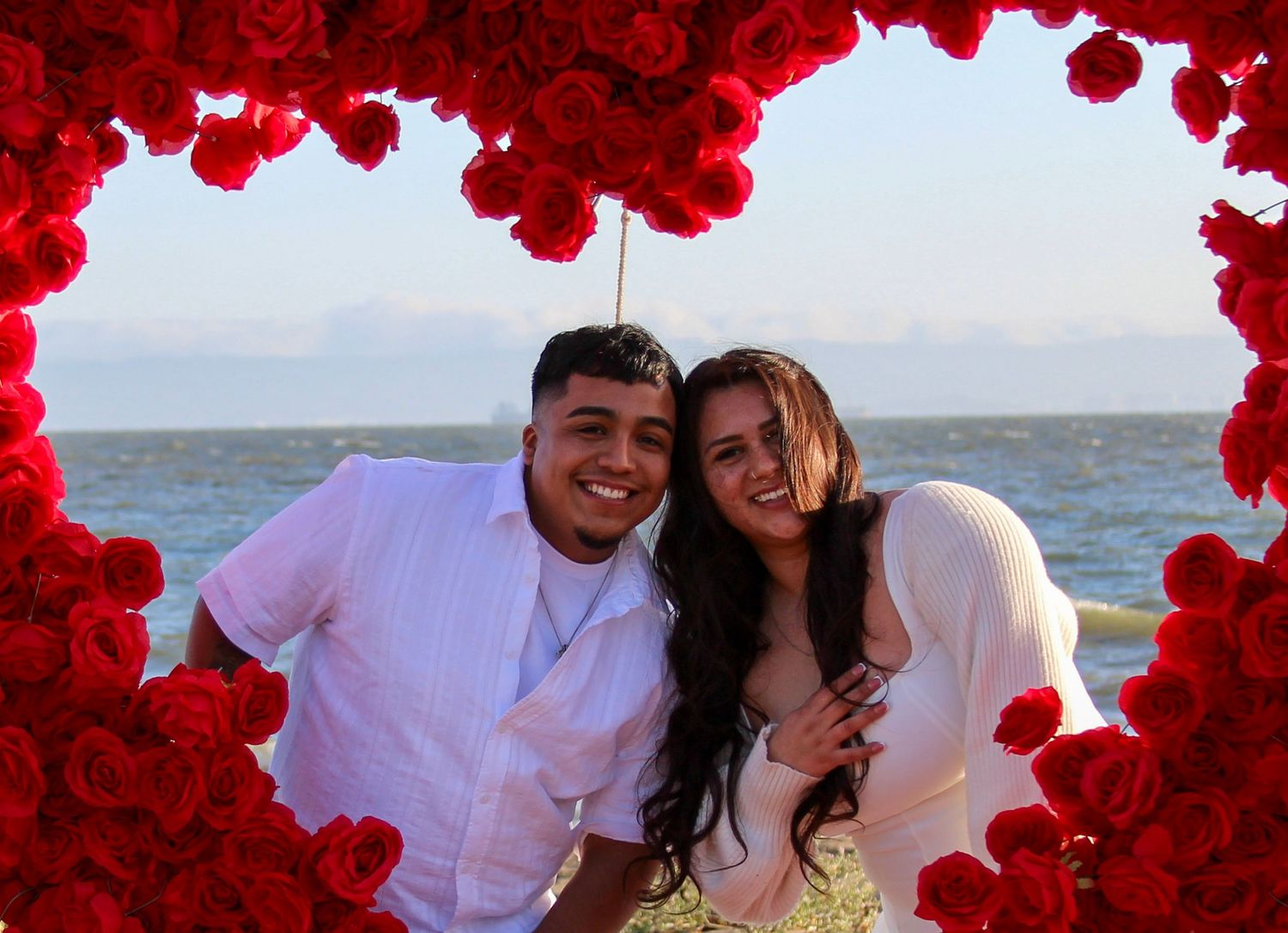Couple smiling inside a heart-shaped red rose frame with ocean backdrop.